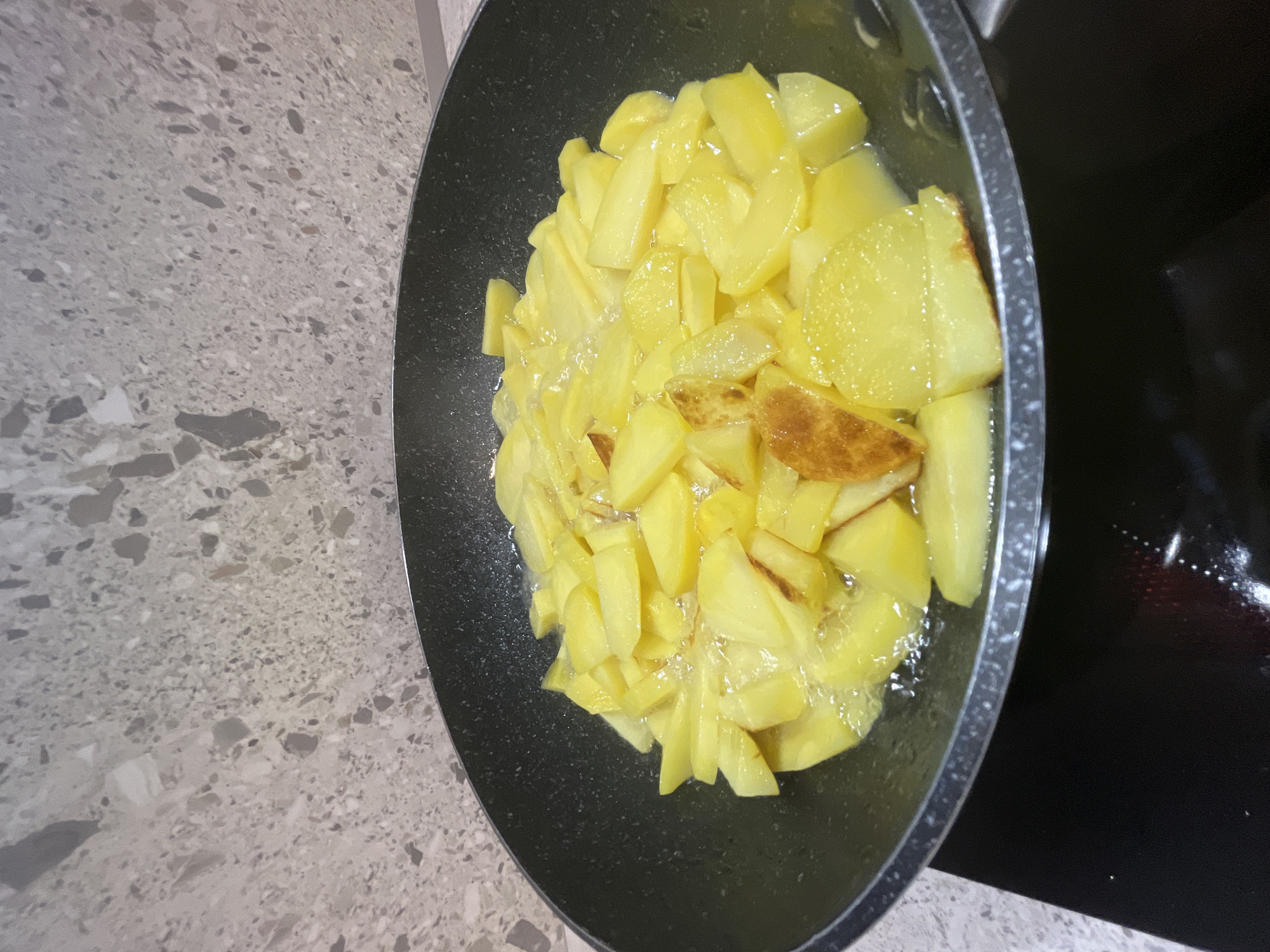 A black frying pan, filled with sliced potatoes being fried in oil, is positioned on a speckled countertop. The potatoes are a vibrant yellow, with some pieces showing browning and a crispy texture. Bubbles from the hot oil are visible around the potatoes, indicating they are actively cooking. The pan's rim is metallic and slightly raised. To the left of the pan, the countertop has a light gray base with a pattern of various sized gray and white speckles. To the right of the pan, a dark surface, likely a stovetop, is visible. The lighting suggests it is daytime. No people are visible in the image.