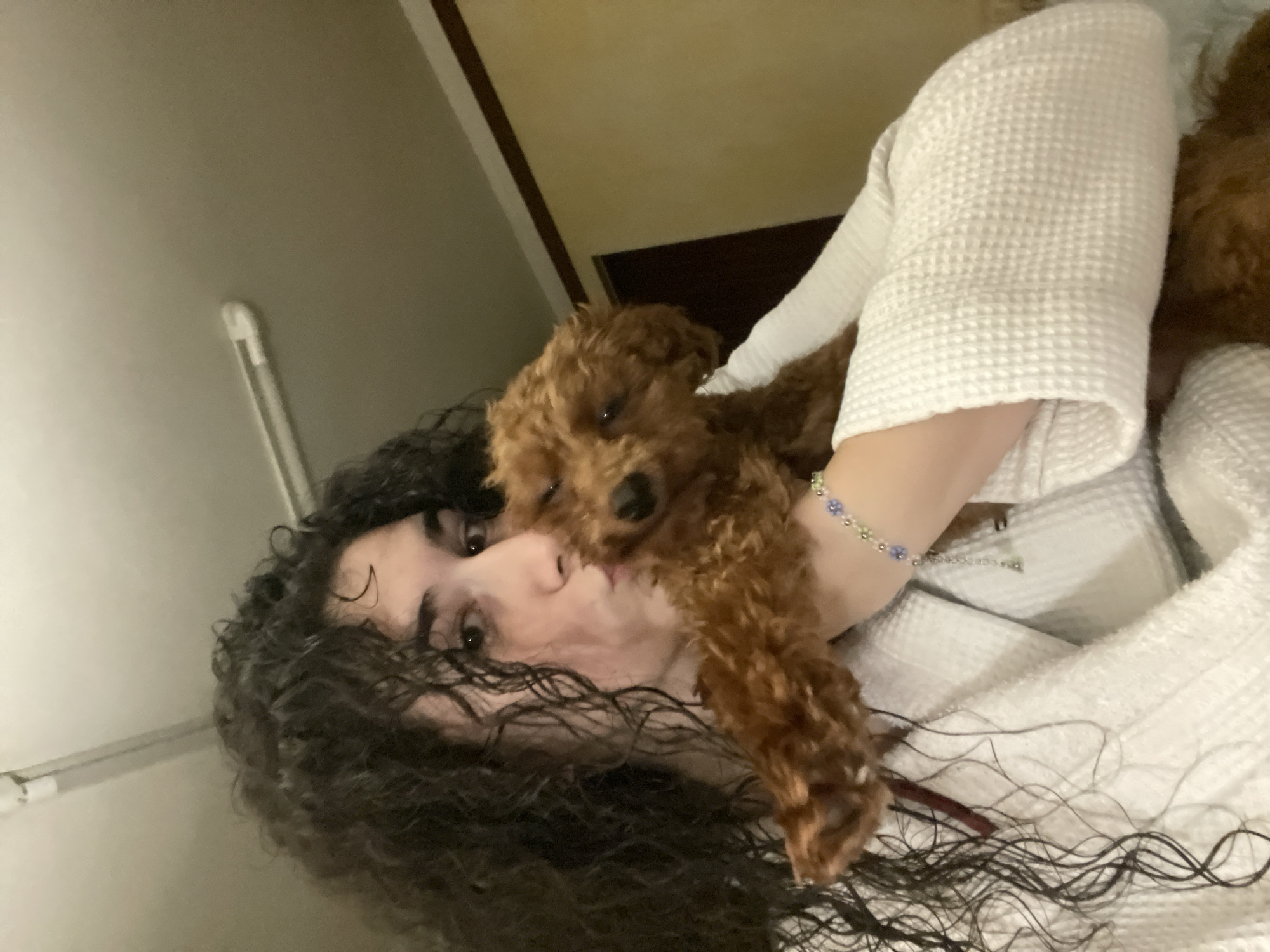 A woman with dark curly hair is holding a small, brown poodle. The woman is wearing a white, textured robe. She is looking at the camera with a neutral expression. The dog is facing forward, also looking at the camera. They appear to be indoors. It is not possible to verify the location is Sofia, Bulgaria, from the visual information provided.