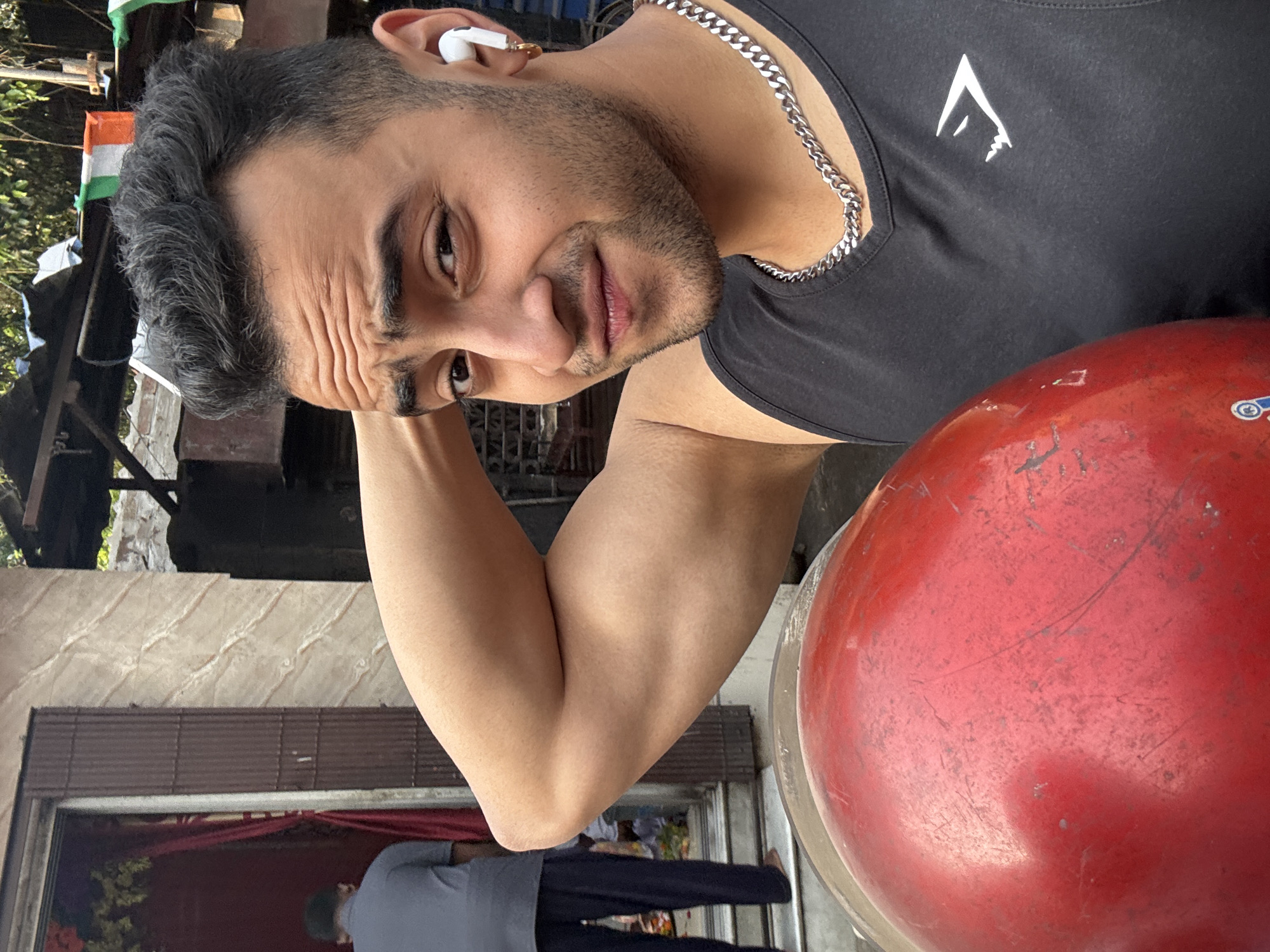 A close-up, slightly angled shot depicts a man with short, dark hair, a trimmed beard, and an earbud in his left ear. He is wearing a black tank top with a white logo and a silver chain necklace. His right arm is raised and flexed, showing a defined bicep. A large, worn, red object, possibly a helmet, is positioned near him.
Behind the man, an exterior scene unfolds. A doorway with a red curtain partially drawn is visible. A person wearing a grey shirt and dark pants stands on steps leading up to the doorway. The architectural style suggests a residential building in an urban setting. Further in the background, there is a view of trees and an Indian flag. The environment appears to be in Kolkata, India, as indicated by the architectural features and the presence of the Indian flag.