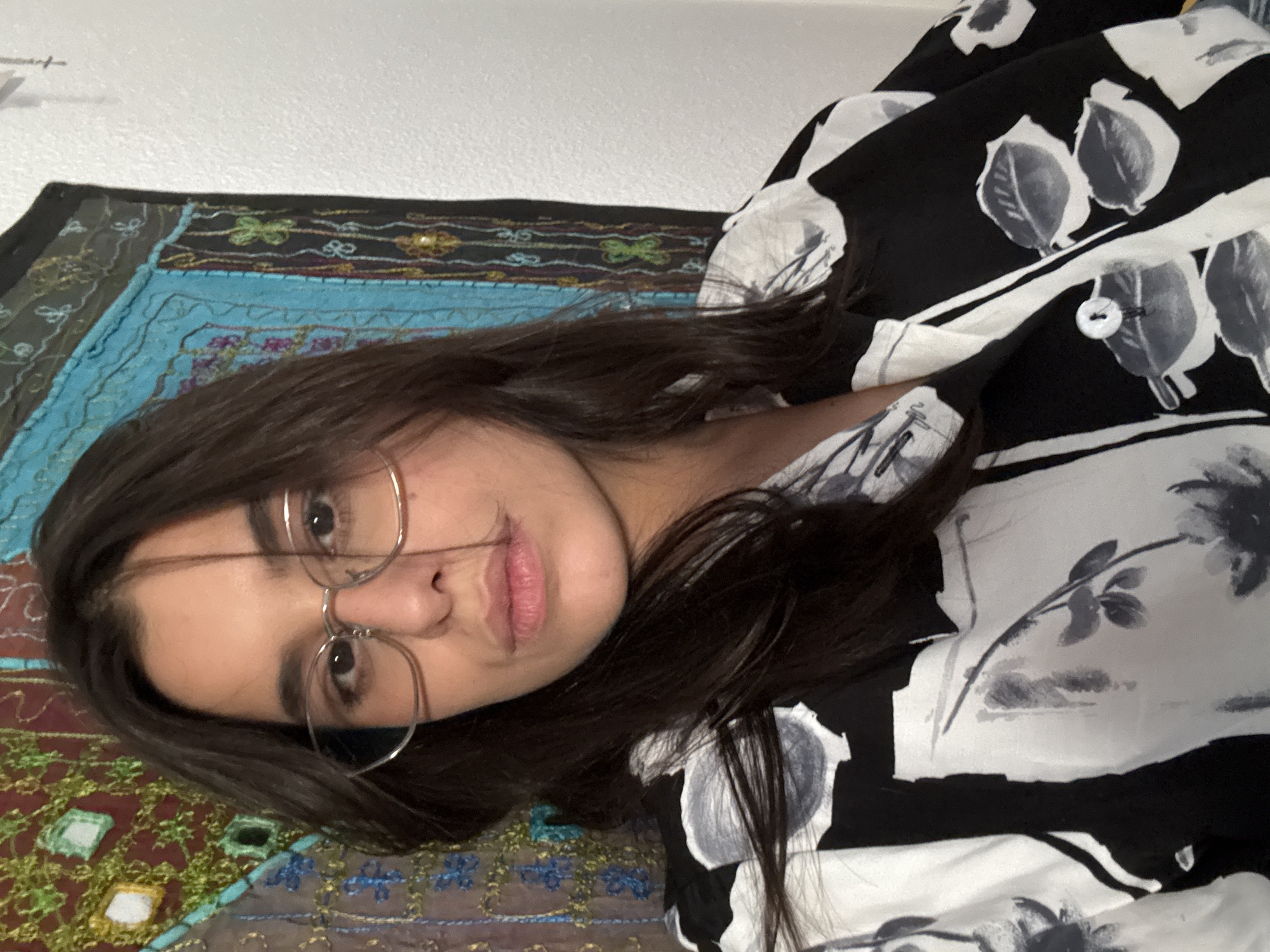 The image shows a close-up of a young woman wearing glasses and a black and white floral print shirt. She has long, dark hair and is looking directly at the camera with a neutral expression. The background features a tapestry with colorful patterns and a white wall. The woman is positioned in the foreground, with the tapestry and wall behind her. There is no indication of the location being in Las Rozas de Madrid, Spain.