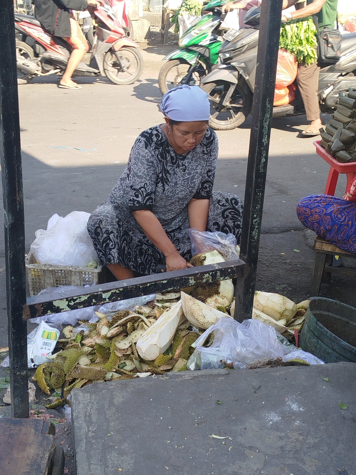 In the city of Larangan, Indonesia, a woman wearing a blue headscarf and a grey patterned dress is seated on the ground, surrounded by discarded durian rinds. She is in the process of preparing durian, with a knife in her right hand and a durian on a plastic bag in front of her. To her left, a large bag of ice is visible in a basket. In the background, a person rides a red motorcycle, and another person wearing a green shirt and brown pants stands near a green and black motorcycle. Another person in a blue and orange patterned sarong sits on a stool to the right. The scene appears to be a street market or roadside stall, with various items for sale, including more durian and packaged goods. The ground is paved with asphalt.