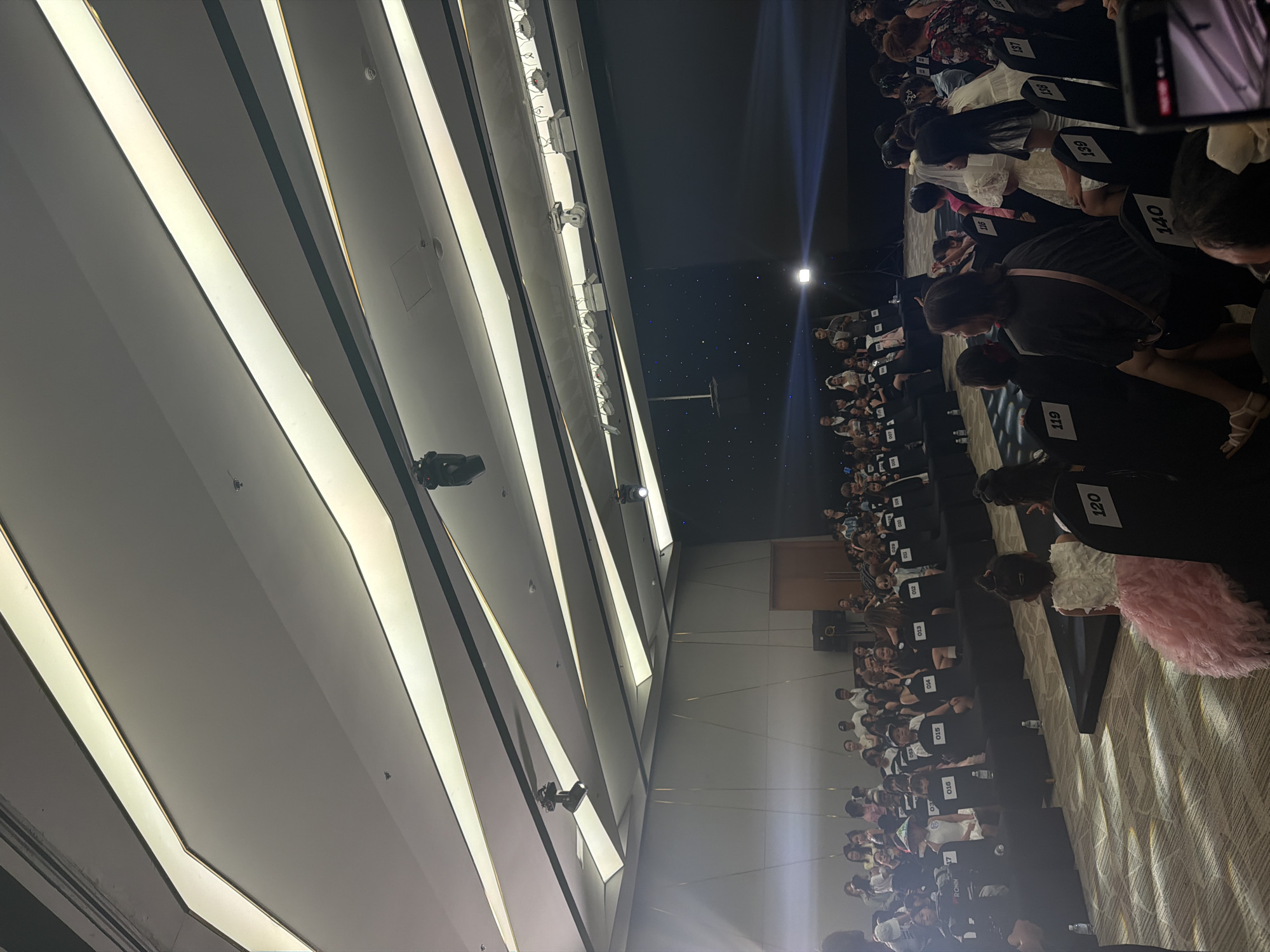 This image captures an indoor event within a large hall or ballroom in Nha Trang Ward, Vietnam. The overhead ceiling is architecturally complex, featuring a series of white, multi-faceted panels integrated with linear light fixtures, some of which are illuminated, and bordered by thin gold-toned lines. Darker, intersecting strips traverse the ceiling, from which several black, cylindrical spotlights are suspended. Various utility features like smoke detectors are also visible on the ceiling.
The left wall presents a light-toned, possibly paneled surface. The rear wall on the right half of the image is dominated by a large, dark screen or backdrop, which displays a pattern resembling a starry night sky. A bright, focused spotlight illuminates the central area of this dark backdrop. A wooden door is positioned on the far right of this rear wall, flanked by visible sound equipment (speakers). The floor is covered with a patterned carpet in light and dark tones.
Multiple rows of black chairs are arranged on the carpeted floor. Many chairs are equipped with white numerical labels (e.g., 007, 014, 120, 140), indicating participant or seating assignments. In the immediate foreground, facing away from the camera, a young girl in a white top and a voluminous pink ruffled skirt occupies chair number 120. Adjacent to her, a partially obscured individual sits in chair 119. To the right, an adult female with dark hair, wearing a dark shirt and a light-colored shoulder bag, occupies chair 140, also facing away from the viewer. A smartphone, held by an unseen person, is visible in the lower right corner of the frame, capturing the scene.
Further into the middle ground, numerous individuals, primarily children and young adults, are seated in the numbered chairs. Noteworthy participants include a child wearing a wide-brimmed hat (chair 017) and another in a white dress (chair 139). A "RONIN" camera stabilizer is positioned on a chair next to participant 017. Small plastic water bottles are visible on the floor beside several chairs. In the background, in front of the dark, starry backdrop, a male adult in a gray shirt is standing, holding a small object, likely a microphone or script, illuminated by a prominent spotlight from the left. Other individuals are observed standing and sitting behind the primary rows of chairs.