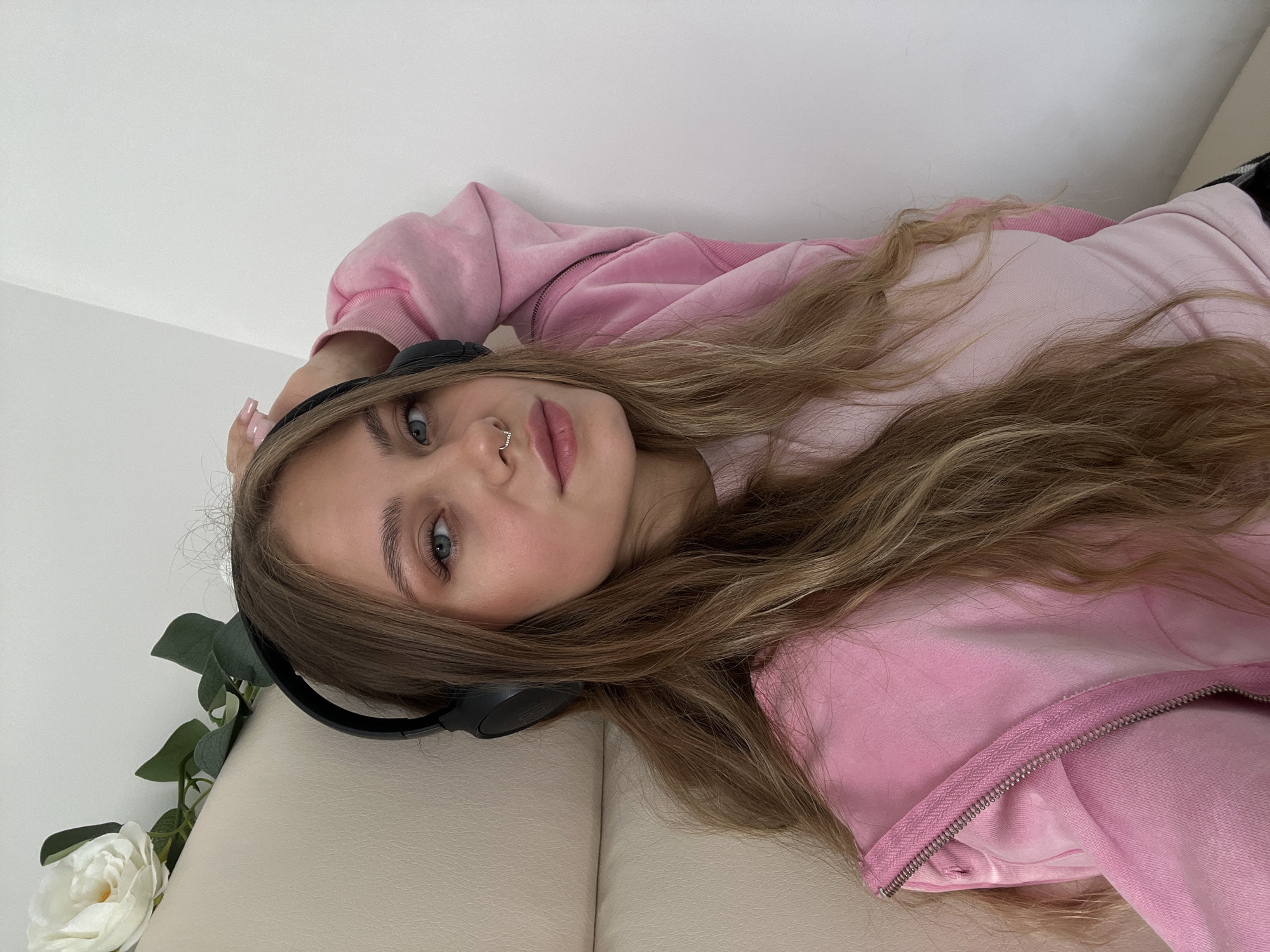 A young woman with long, wavy blonde hair and blue eyes is seated, wearing a pink hoodie and headphones. She has a nose ring and her left arm is raised behind her head. A white artificial rose with green leaves is visible behind her left ear. The background consists of a light-colored wall and a cream-colored textured surface, possibly a chair or sofa. The scene appears to be indoors, with no specific city or country identified.
