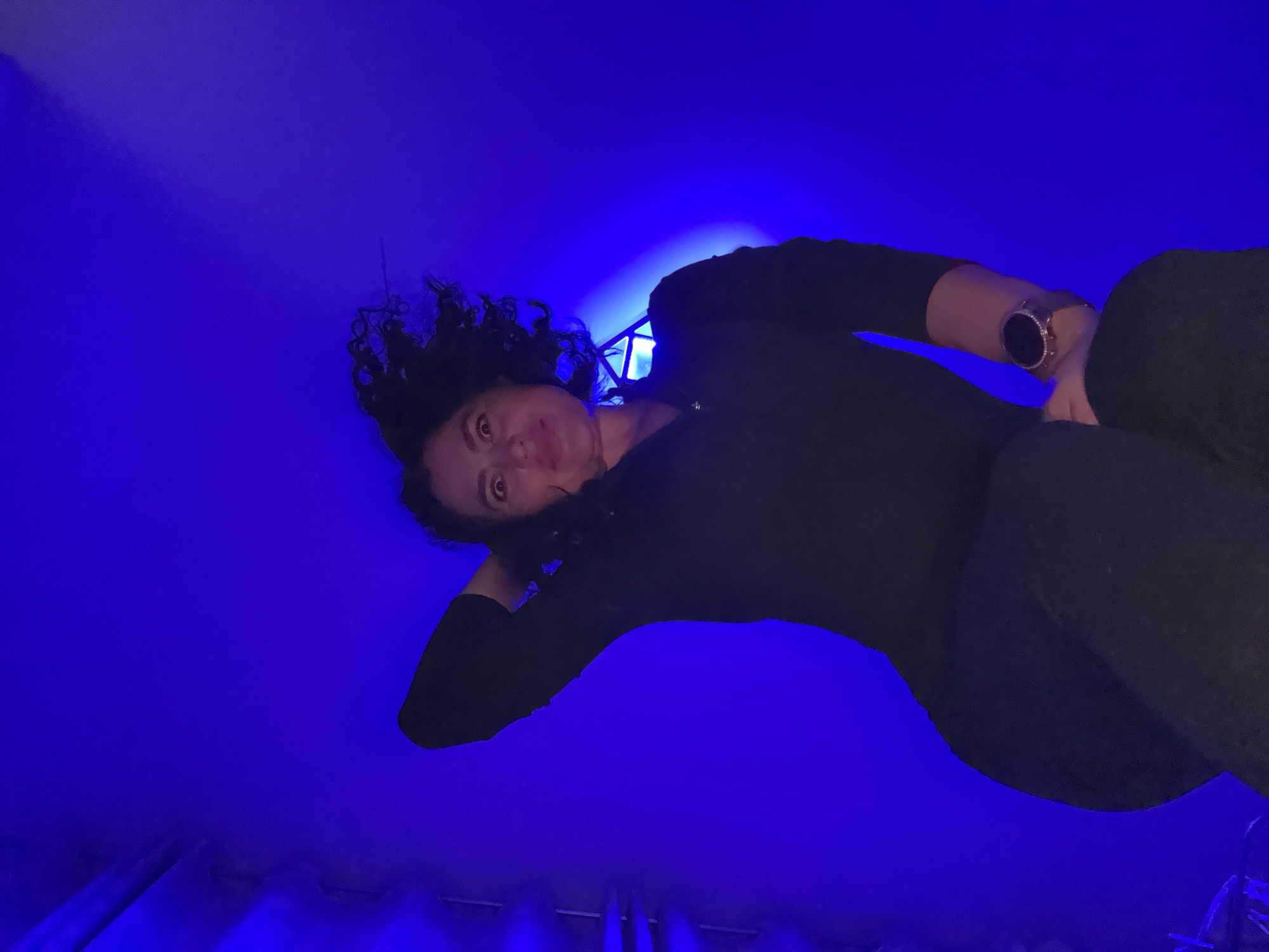 A woman with dark, curly hair is looking down at the camera. She is wearing a dark, long-sleeved shirt and a watch on her left wrist. The background is a bright blue with some lighting fixtures visible at the bottom. The image appears to be taken from a low angle, looking up at the woman. There is no clear indication of the location being in Kauchuk Stantsiya, Russia.