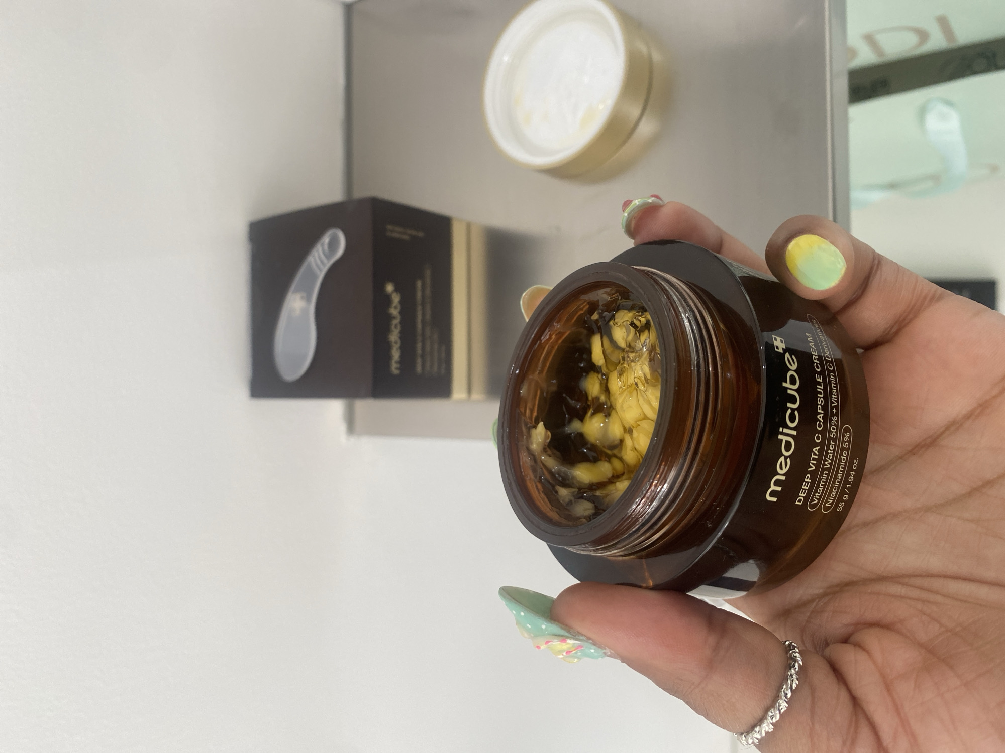 A person's hand, exhibiting a dark skin tone, is prominently featured in a close-up shot, holding an open jar of skincare cream. The thumb's nail is painted with a vibrant yellow and green ombre design, while the ring and pinky fingers display a light green base adorned with white polka dots and delicate floral accents. A silver ring with a twisted rope-like design is worn on one of the fingers.
The product held is a brown, amber-tinted glass jar from the brand "medicube," specifically the "DEEP VITA C CAPSULE CREAM." The jar is filled with a unique formulation consisting of numerous small, irregularly shaped yellow capsules suspended within a clear or light-colored gel. The label on the jar is clearly legible, detailing key ingredients such as "Vitamin Water 50% + Vitamin C Derivatives" and "Niacinamide 5%," along with the product weight of "55 g / 1.94 oz." The lid of the cream jar, with its cream-colored interior and golden exterior, is visible in the background, resting on a surface.
To the left, a dark brown "medicube" product box is partially visible, featuring a translucent, curved tool illustration. The setting appears to be indoors, possibly a retail environment or a personal display area in London, United Kingdom, characterized by clean white and metallic surfaces. In the blurred background, part of another item with text fragments "DI" and "COL" is discernible. The image is brightly lit, emphasizing the textures and colors of the cream and the person's hand.