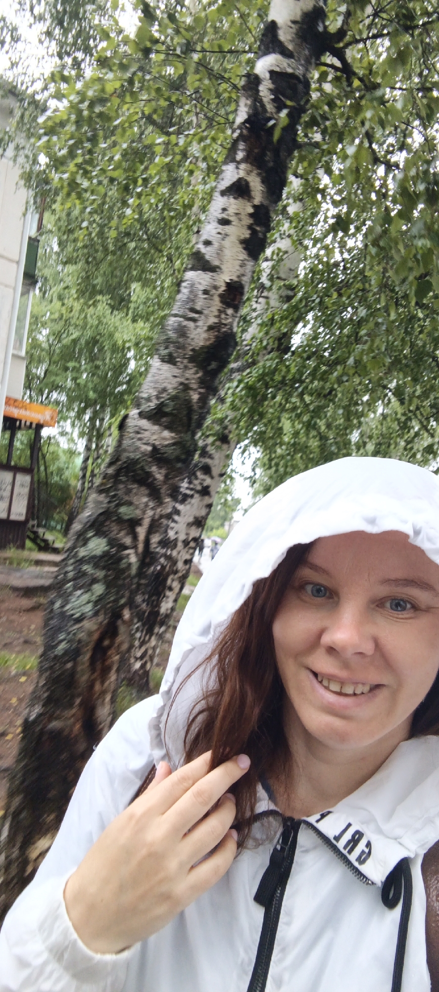 A woman with brown hair and blue eyes smiles and touches her hair while wearing a white hooded jacket. She is standing in front of a birch tree with mottled bark. Green leaves from the tree and other foliage are visible in the background, along with a building and a sign. The scene appears to be outdoors, possibly in a park or residential area, given the presence of the building and the natural elements.