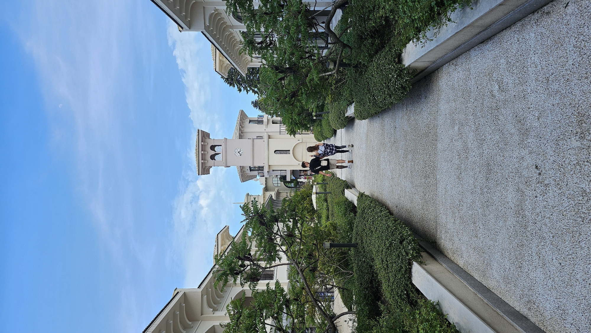 The image shows a couple walking down a paved pathway lined with greenery and buildings. The pathway is made of a light gray, speckled material and is bordered by concrete edges. On either side of the path, there are dense bushes and trees. The buildings are a light color with arched windows and decorative details. In the background, there is a clock tower. The sky is blue with some clouds. The couple consists of a man wearing a black shirt and beige shorts, and a woman wearing a plaid shirt and dark shorts. They are walking side-by-side. The location is likely in Varna, Bulgaria, given the architectural style of the buildings and the surrounding environment.