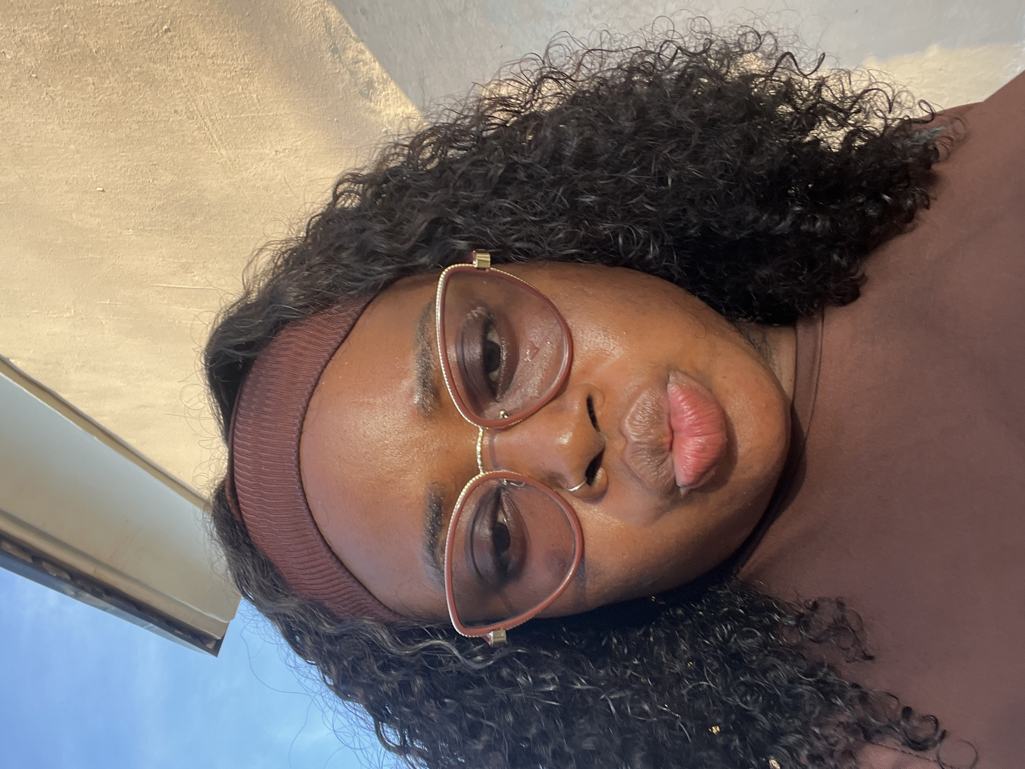A selfie of a Black woman with curly dark hair and a brown headband is taken outdoors, likely in Abuja, Nigeria. She is wearing large, light brown-tinted glasses with gold accents and a brown top. Her lips are pursed, and she is looking at the camera. Behind her, a lightly textured cream-colored wall casts a shadow, and the blue sky is visible at the top left of the frame. A portion of a building's overhang is also visible in the upper left. The lighting suggests it is either early morning or late afternoon.