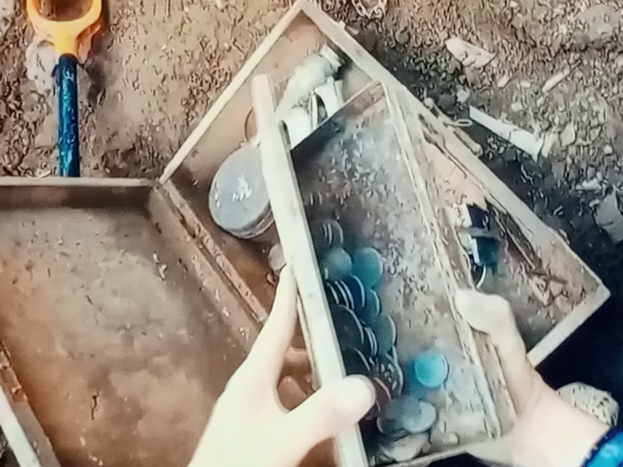 In an outdoor scene with dirt and gravel ground, a person's hands are seen holding an open wooden box. The box contains several round, metallic objects that appear to be coins, some of which have a blueish hue. A blue and orange trowel is visible in the upper left corner, partially embedded in the ground. The scene is viewed from a high angle.
