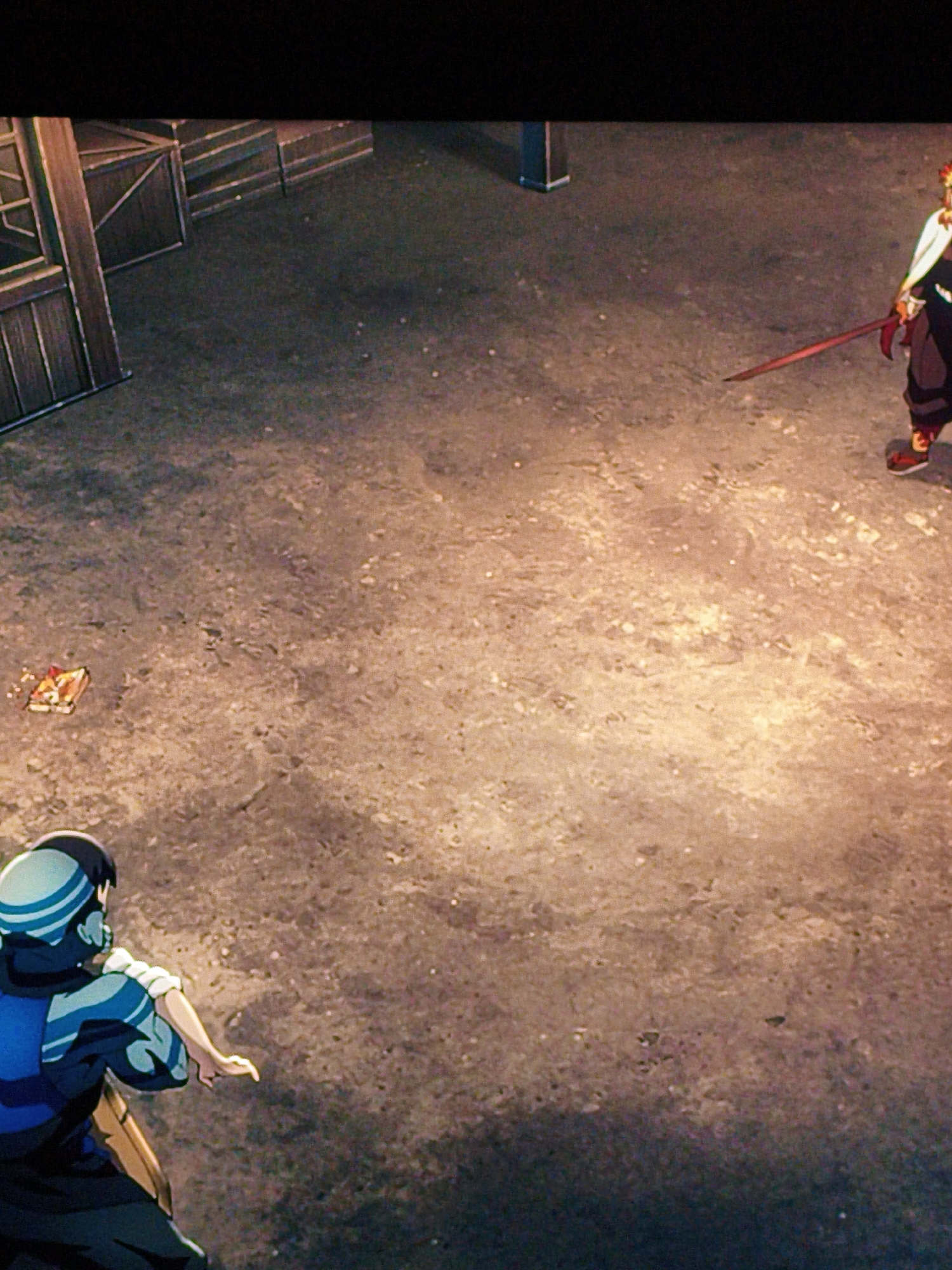 The image depicts an animated scene, likely from a video game or anime, set in an outdoor, possibly urban, environment. In the foreground, a character with a blue and white outfit and a blue head covering is shown from behind, facing to the right with their arm extended. In the mid-ground, a small, rectangular object with red and yellow markings lies on the dusty, concrete ground. Further in the background, another character, appearing to be male, is visible from the side. This character has red and yellow hair and is holding a red sword horizontally. The environment features a wooden structure resembling a building or fence on the left side of the frame. The lighting suggests either daytime with artificial light sources or nighttime with significant illumination. There is no discernible information within the image to suggest the location is Vladivostok, Russia.