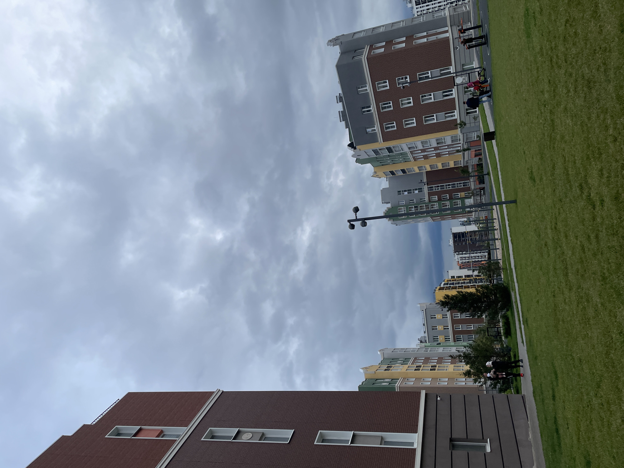 The image depicts a modern residential complex in Pervomaysky, Russia, under a cloudy sky. In the foreground, a well-maintained green lawn leads towards a paved path and a small outdoor exercise area. Several contemporary apartment buildings with colorful facades and numerous windows line the background. The buildings feature a mix of brick and painted surfaces in shades of brown, red, yellow, green, and gray, with distinctive stepped rooflines. A few evergreen trees are scattered throughout the complex. In the mid-ground, a group of people, including adults and at least one child, are visible near a path and some outdoor fitness equipment. One adult is holding a baby. Another group of individuals is near the exercise equipment. The scene suggests a residential area with amenities for its inhabitants.