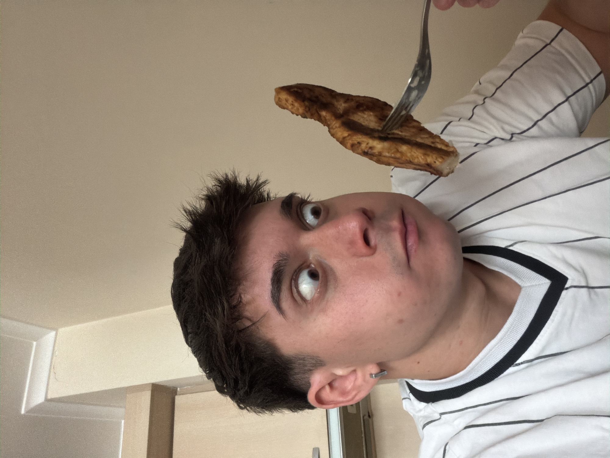 A low-angle, close-up shot shows a young man with short, dark hair and light skin. His eyes are directed upwards towards a piece of food, likely bread or toast, being held above his head with a fork. He is wearing a white t-shirt with black stripes. The background is a light beige wall and a section of what appears to be a kitchen cabinet. There is no direct evidence within the image to confirm the location as Sofia, Bulgaria.