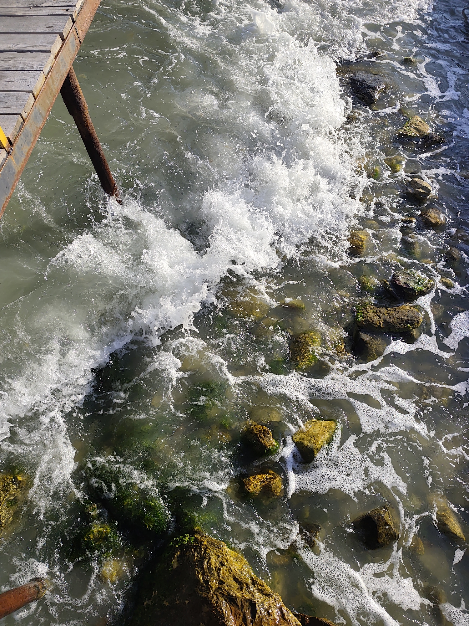 This media file captures a dynamic scene of waves crashing against a rocky shore next to a wooden pier. The water is turbulent, with white foam and spray indicating significant wave action. The shore is littered with various-sized rocks, many of which are partially submerged and covered in green algae. A weathered wooden pier extends into the frame from the left, supported by a rusty metal pole that dips into the churning water. The scene suggests a coastline with a rougher sea, possibly near a dock or harbor. The lighting indicates it is daytime, with sunlight illuminating the water and rocks, creating highlights and shadows. There is no visible text in the media file.