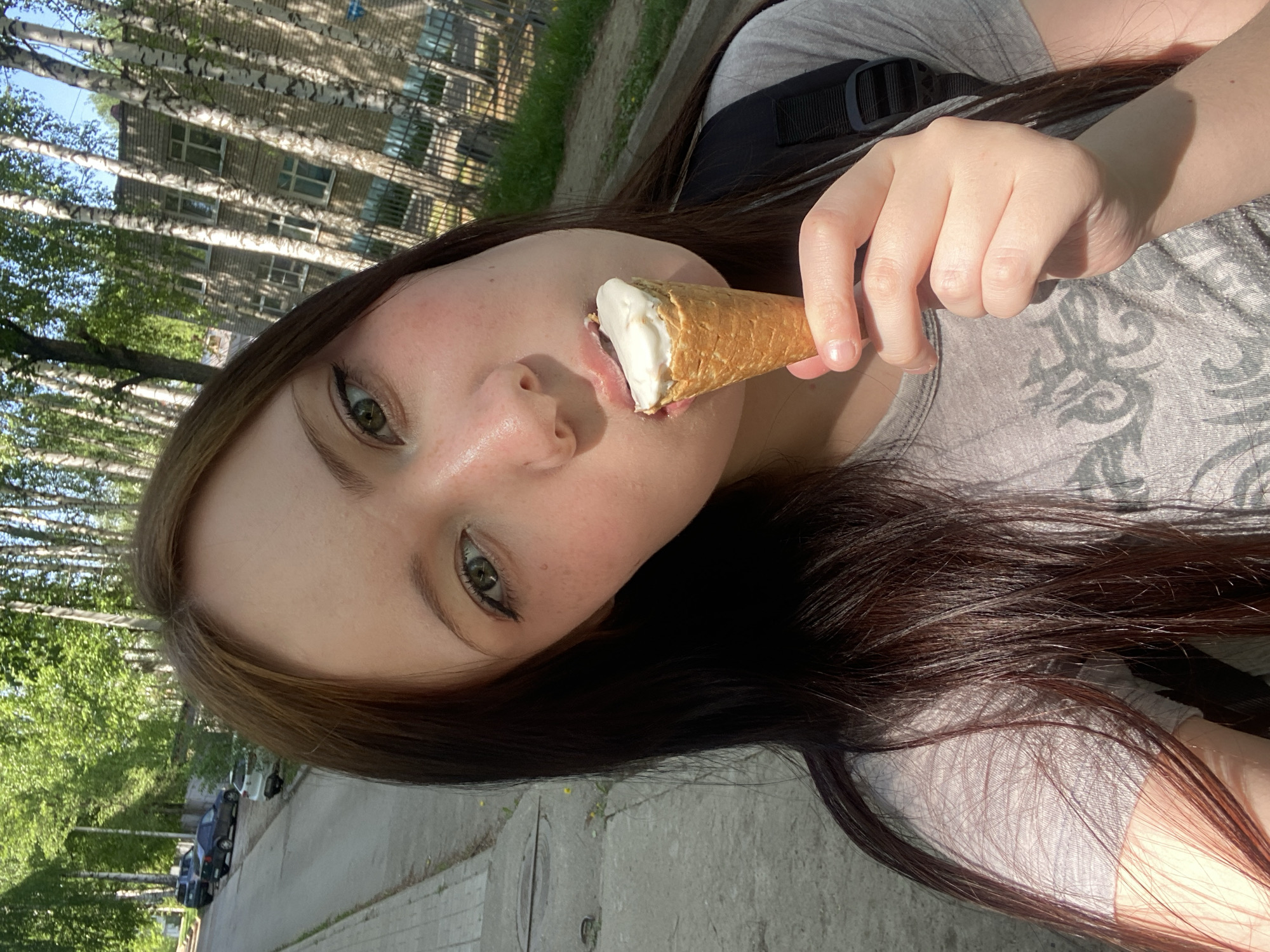 A young woman is holding an ice cream cone to her mouth in an outdoor setting. She has long brown hair with lighter brown streaks and green eyes. She is wearing a gray t-shirt and a black backpack. The background shows a street with trees lining the sidewalk, and a building with many windows is visible on the left. Several cars are parked along the street. The location appears to be a residential area within the city of Unknown, Unknown.