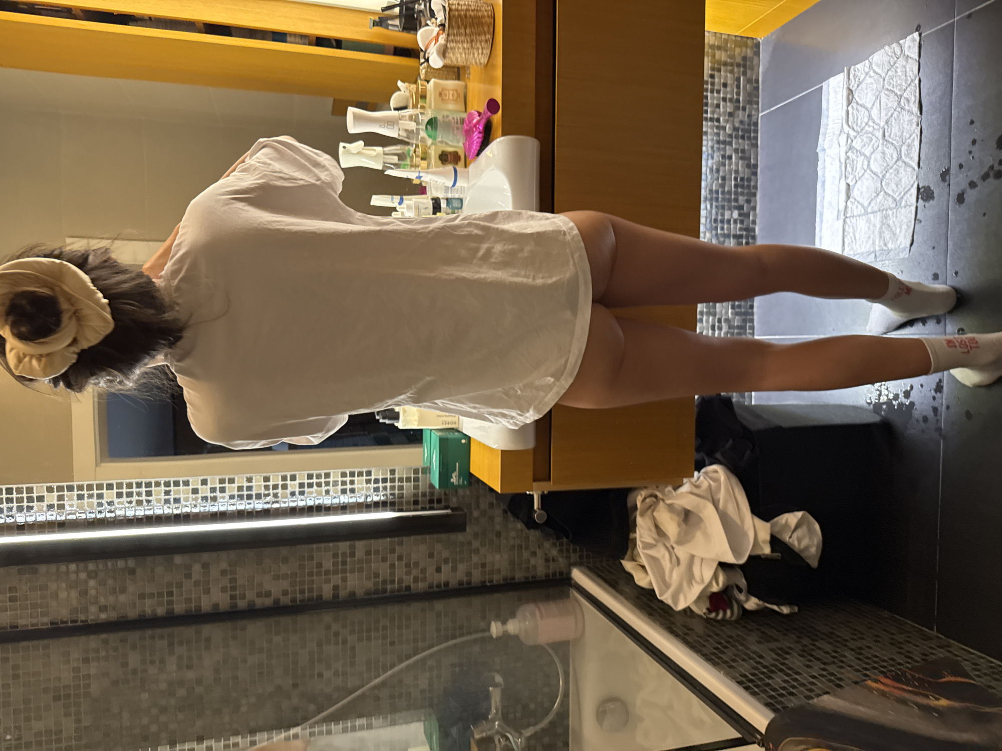 The scene takes place in an indoor bathroom at an unspecified location in the city of Unknown, Unknown. A person is depicted from the back, standing in front of a light-colored wooden bathroom vanity with a white, rectangular basin. The individual has dark hair pulled back with a light-colored scrunchie and is wearing a loose-fitting white short-sleeved T-shirt and white socks, revealing their buttocks and upper thighs as no lower garments are visible.
The vanity countertop is cluttered with numerous personal care products, including bottles, pump dispensers, a green box labeled "MORPHE," a pink comb, and a small woven basket filled with items. A mirror is present above the vanity, flanked by vertical strip lights. To the left of the vanity, a shower area with a glass screen and a white bathtub is visible. The walls in this section are tiled with small, square mosaic tiles in varying shades of grey and black. A light pink pump bottle is on the edge of the bathtub.
The floor is dark grey tiled, featuring a white quilted mat or towel placed in front of the vanity. Numerous water droplets are scattered across the dark floor tiles, particularly around the person's feet and the mat. To the left of the vanity, a dark-colored stool or ottoman holds a pile of crumpled clothing, predominantly white, with some darker items.