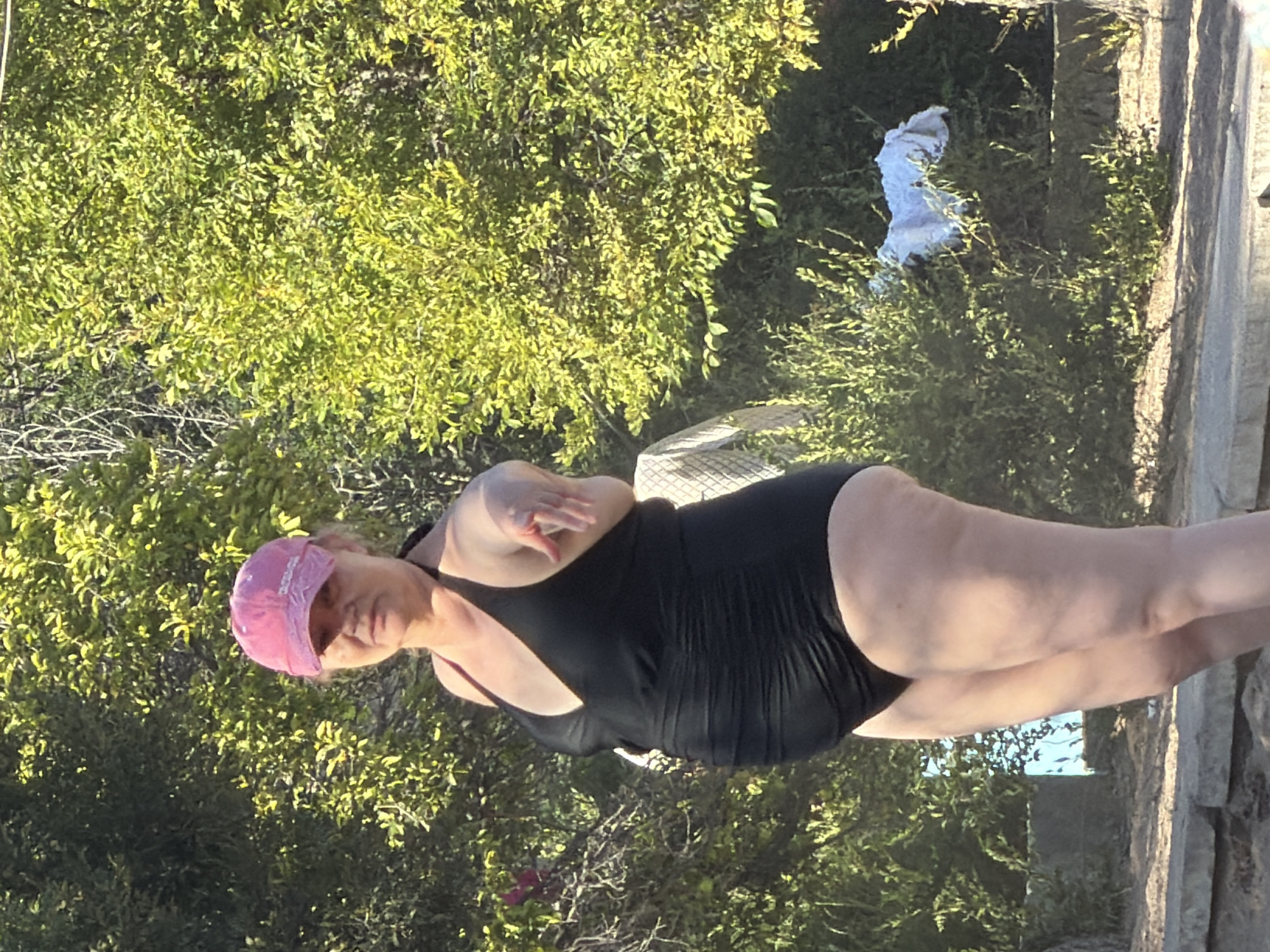 A woman wearing a pink baseball cap and a black one-piece swimsuit stands outdoors. She is facing the camera and pointing with her right hand. She has fair skin and her hair is pulled back. Behind her, there is lush green foliage. The ground in front of her is covered in dirt and stone.
