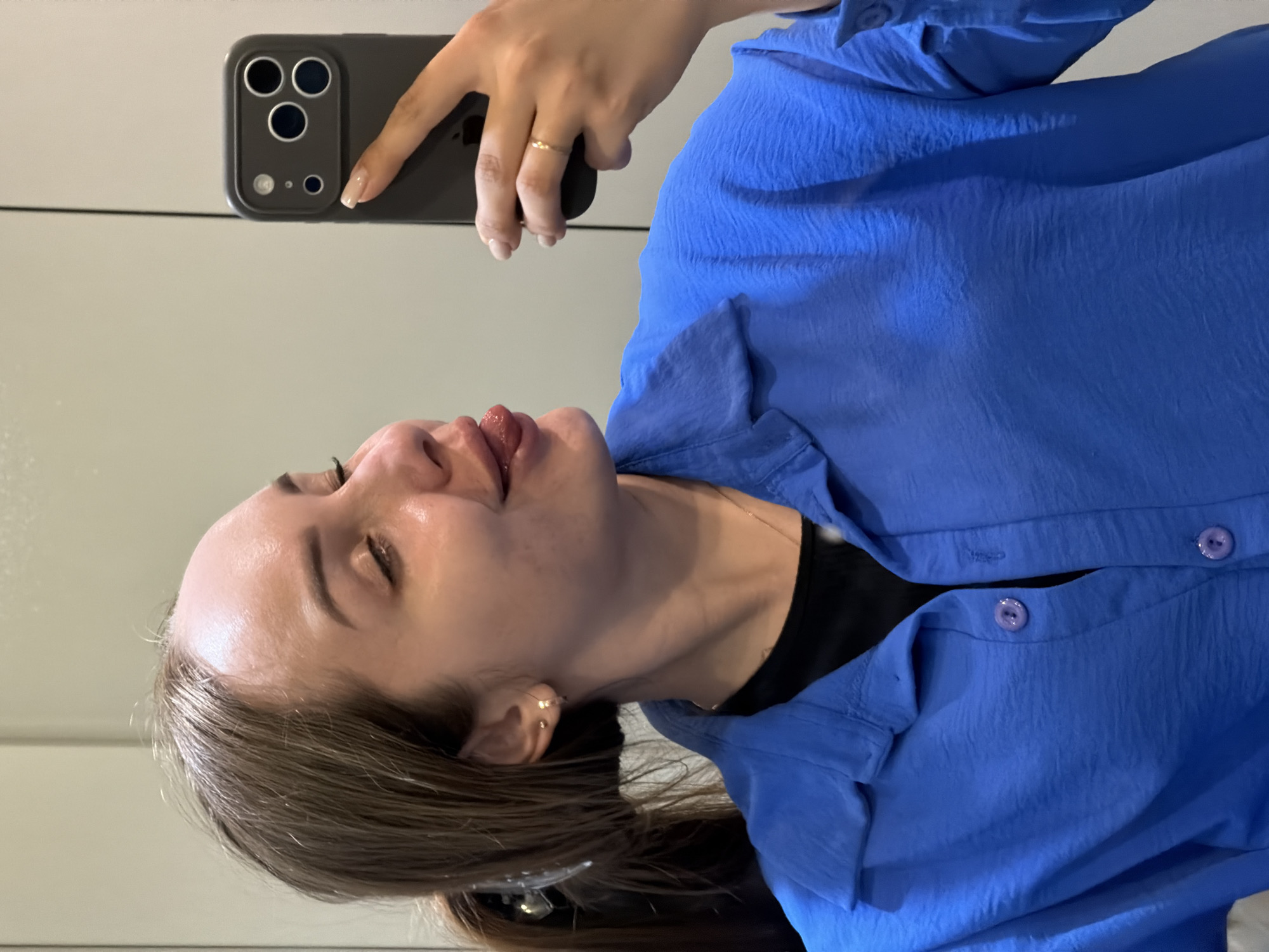A woman is taking a selfie in what appears to be a bathroom. She is wearing a blue button-down shirt over a black top. She is holding a smartphone with a dark gray case and multiple camera lenses. The location is indeterminate, there is nothing in the image that would indicate this image was taken in Chaikovsky, Russia.