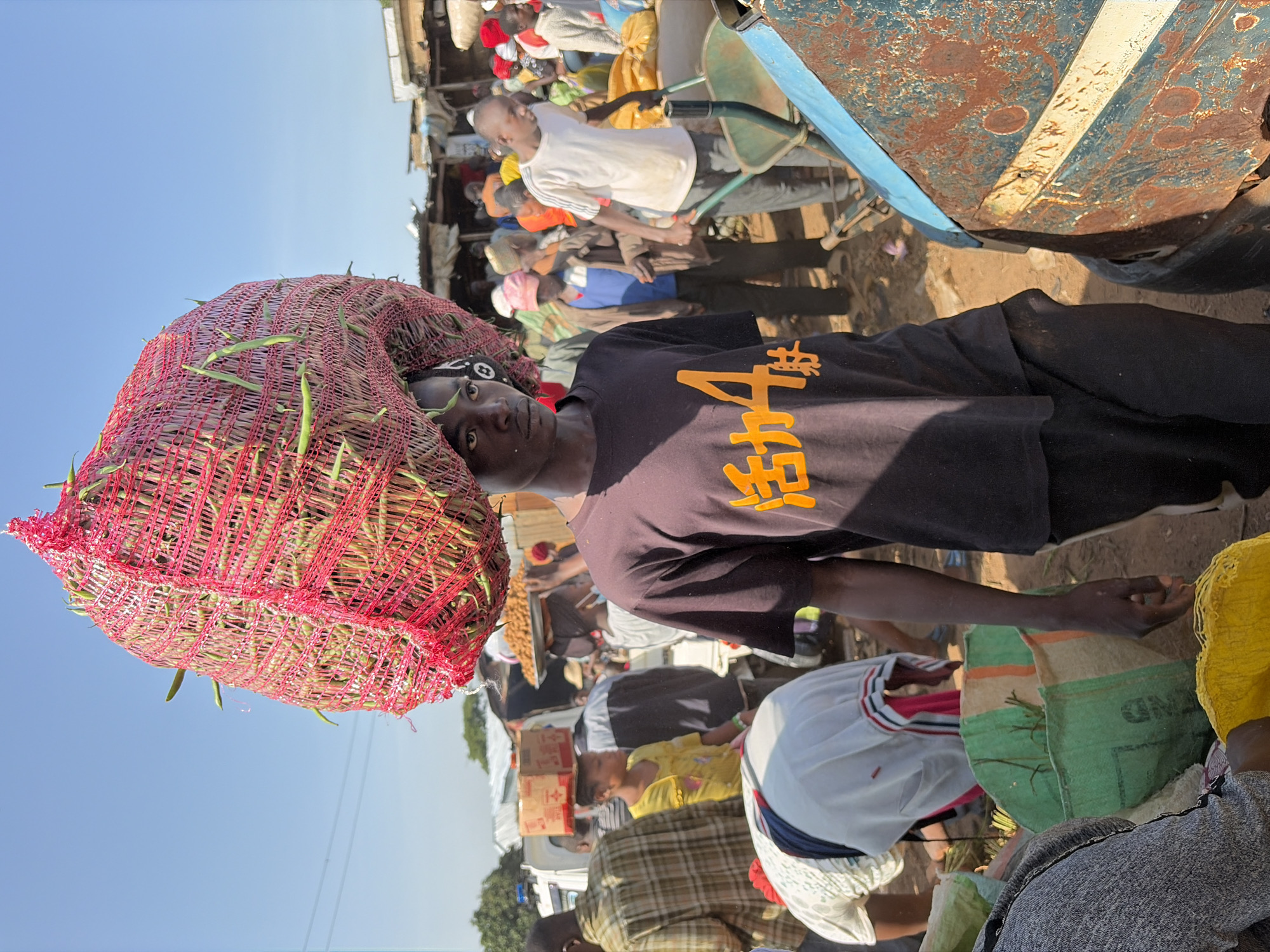 A young man is depicted from the chest up, carrying a large net bag filled with green beans on his head. The bag is a vibrant red mesh, and the beans are clearly visible protruding from it. He is wearing a dark t-shirt with orange lettering that appears to be in an Asian script. The man is in a crowded outdoor market, with many other people in the background. The market stalls are made of corrugated metal and wood. In the background, a rusty blue vehicle is visible, with part of its front fender and hood taking up the right side of the frame. The ground is dusty and unpaved. The sky is a clear blue. The scene is likely in the city of Gura Top, Nigeria, based on the context provided.