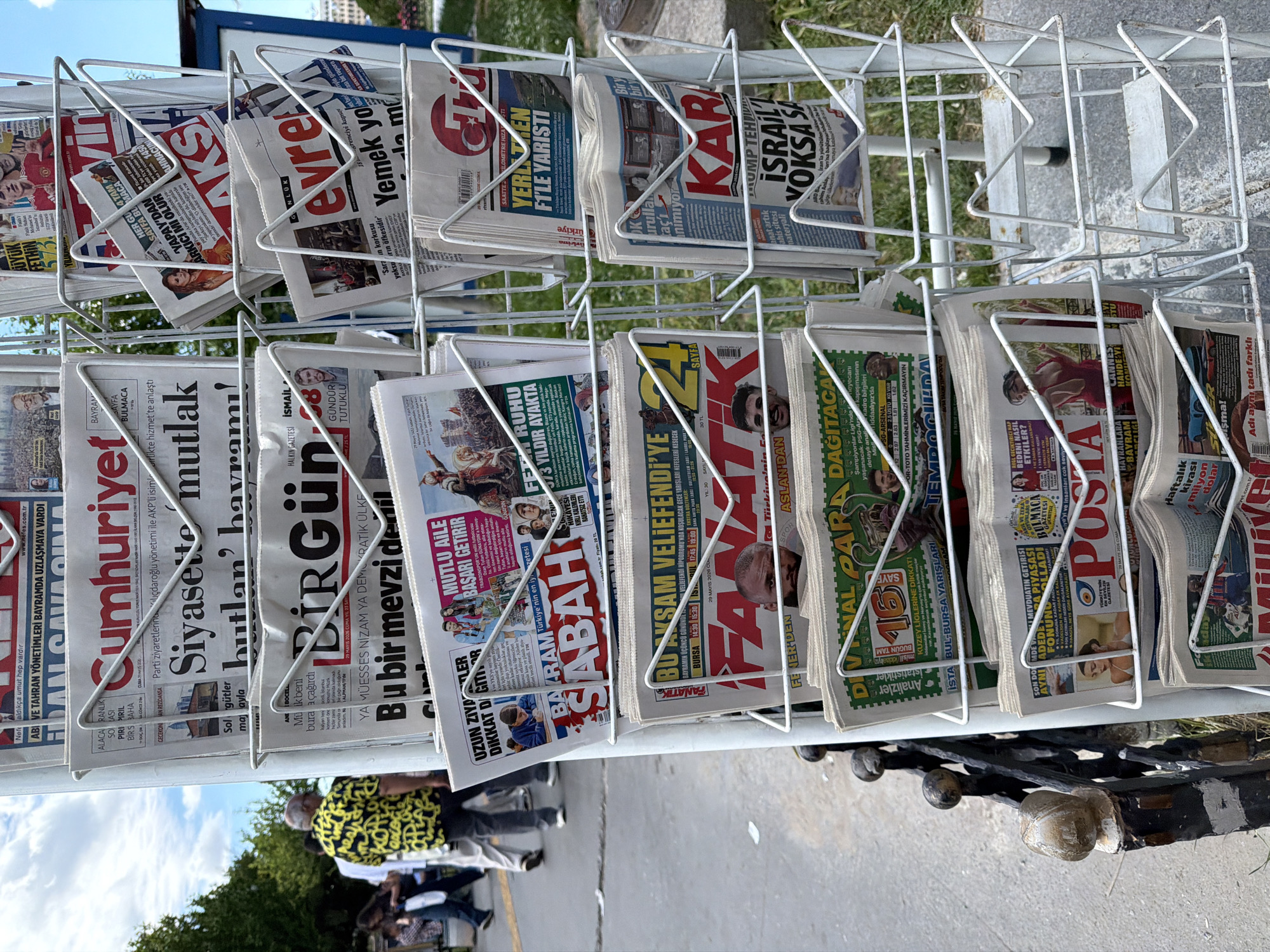 This photo depicts a street-side newspaper stand in Kadıköy, Turkey. The stand is a white wire rack displaying a variety of Turkish newspapers, including Cumhuriyet, BirGün, Sabah, Fanatik, Posta, Karar, and Evre, among others. The papers are stacked and partially visible.
In the background, a person wearing a yellow and black patterned shirt is visible along with other individuals passing by. The setting appears to be an urban environment with trees and buildings visible in the distance under a clear blue sky. The ground is paved, and the newspaper stand is positioned near the curb.
