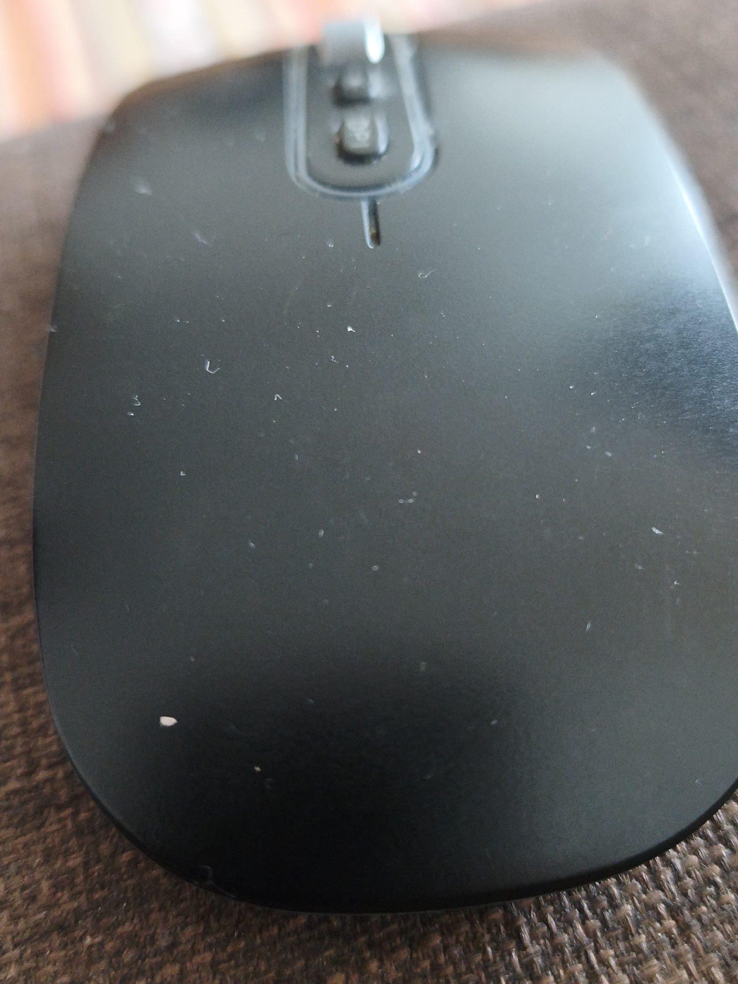 The image shows a close-up, overhead view of a black computer mouse resting on a brown, textured surface. The mouse has a smooth, matte finish and features two buttons and a scroll wheel at the top. Several small white specks, likely dust or lint, are visible on the surface of the mouse. The background is out of focus, with hints of muted colors suggesting an indoor environment. There is no indication of the location being in the city of Duisburg, Germany.