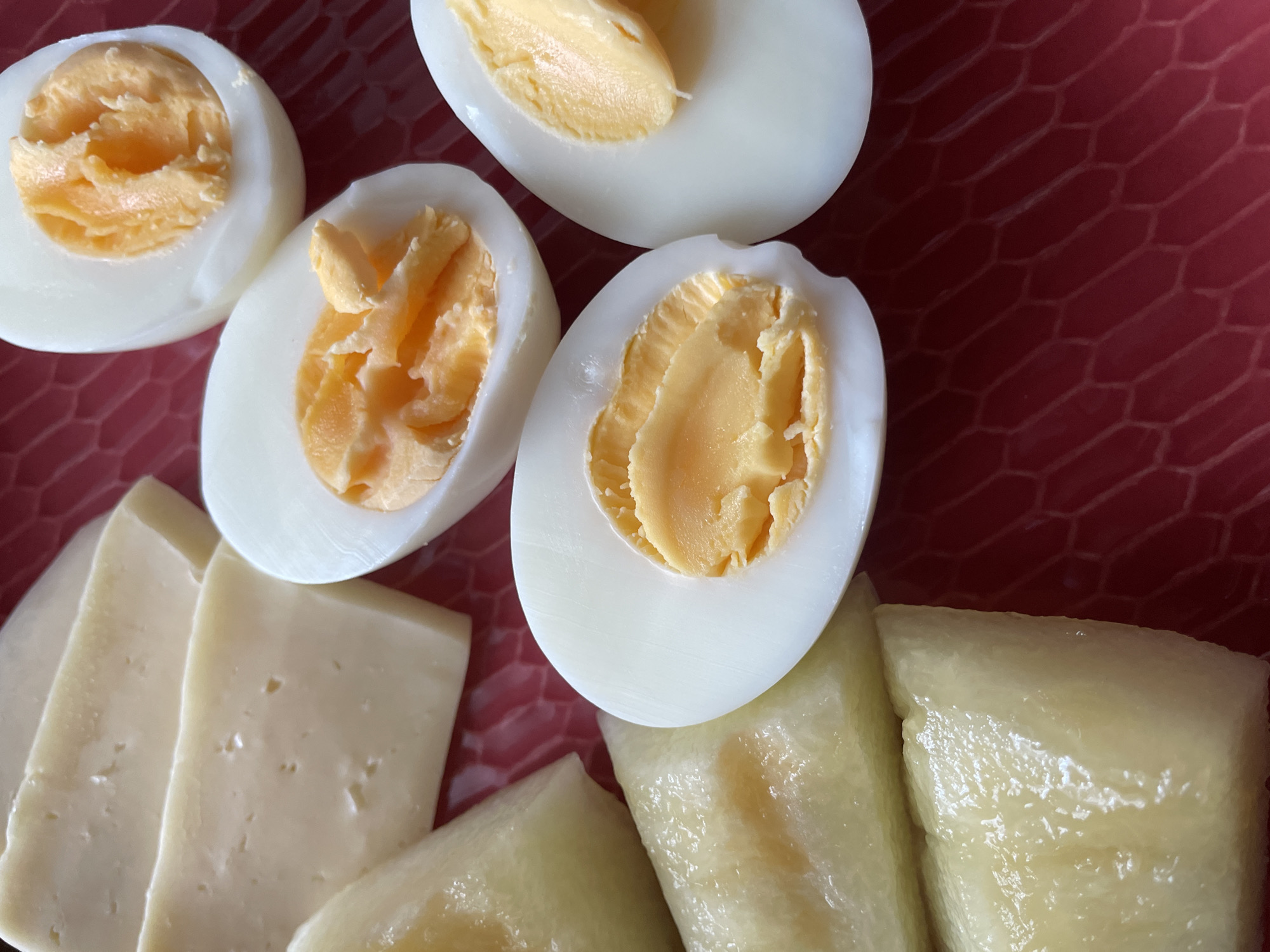 A close-up, overhead shot showcases a red, textured plate filled with slices of cheese, pieces of hard-boiled egg, and some pale, rectangular objects that appear to be cut pieces of cheese or perhaps a pastry. Three halves of hard-boiled eggs are visible, with their bright yellow yolks exposed. The cheese slices are light yellow and have small, irregular holes. The pale rectangular objects are slightly translucent and have a smooth surface. The background is a deep red plate with a honeycomb-like pattern. No people or actions are depicted.