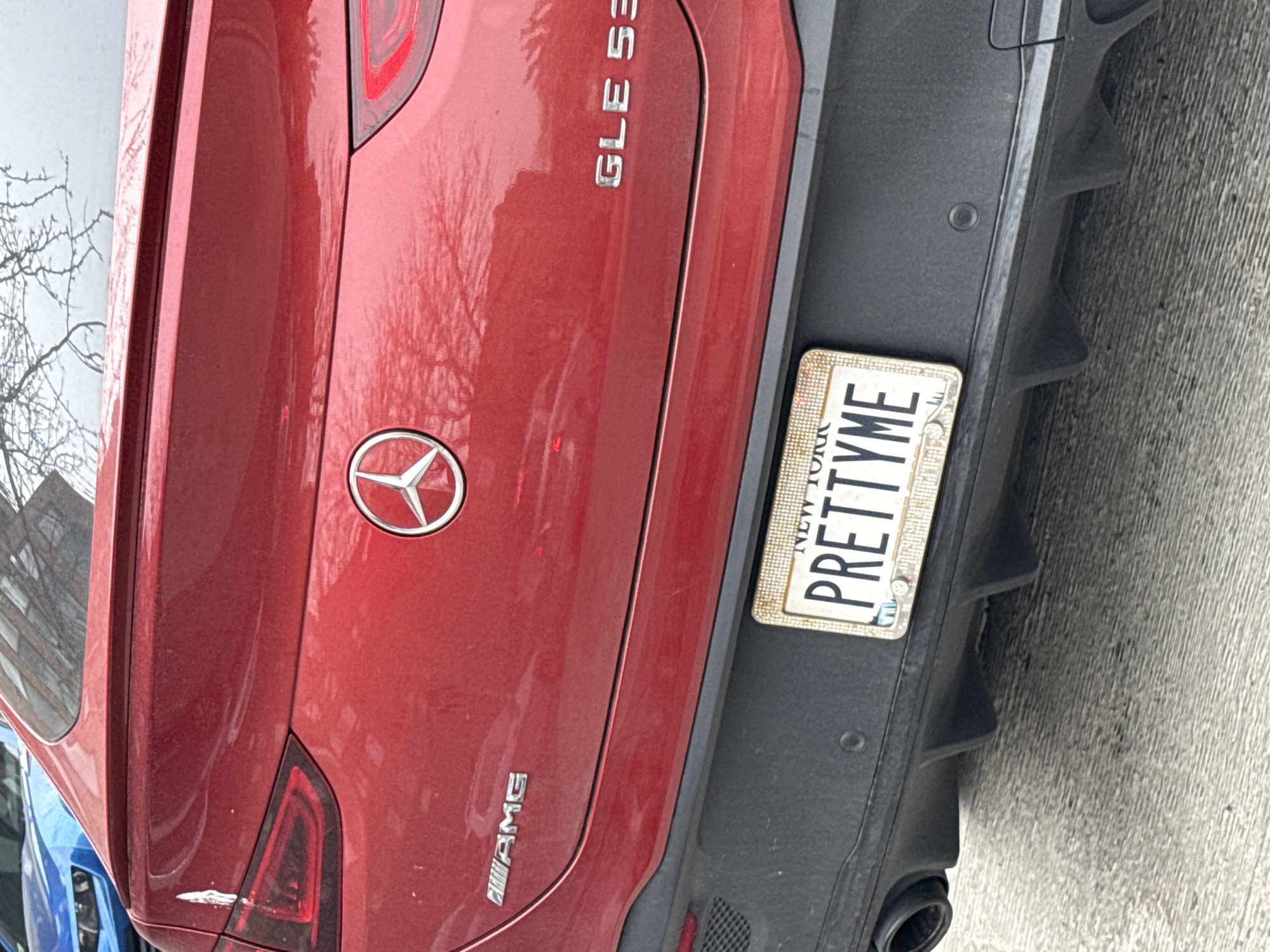 The image shows the rear of a red Mercedes-Benz GLE 53 AMG parked on a street. The car has a New York license plate with the personalized text "PRETTYME". The car's AMG badging and Mercedes-Benz emblem are visible. The car is partially angled relative to the camera. The surroundings include what appears to be part of a building reflecting in the car's surface and a glimpse of a blue car behind it. The background suggests an urban environment.