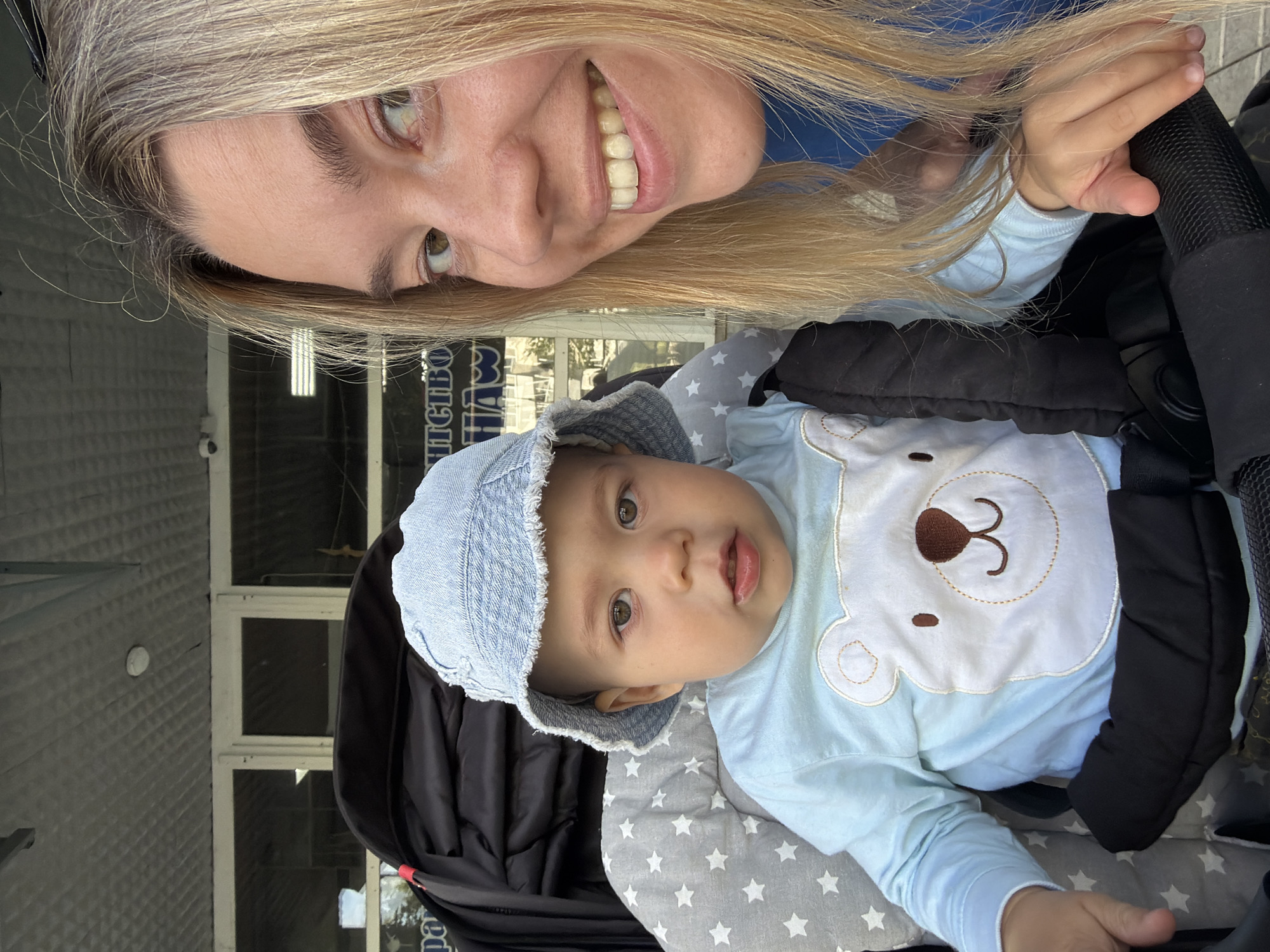 A mother and her baby are in a stroller outdoors, possibly on a sidewalk in front of a building with large windows. The baby, wearing a blue denim hat and a light blue long-sleeved shirt with a white bear applique, is seated in the stroller. The mother, with blonde hair, is smiling and taking a selfie. The background shows a building facade with windows, suggesting an urban setting. There are no specific landmarks that identify the location as Chaikovsky, Russia.
