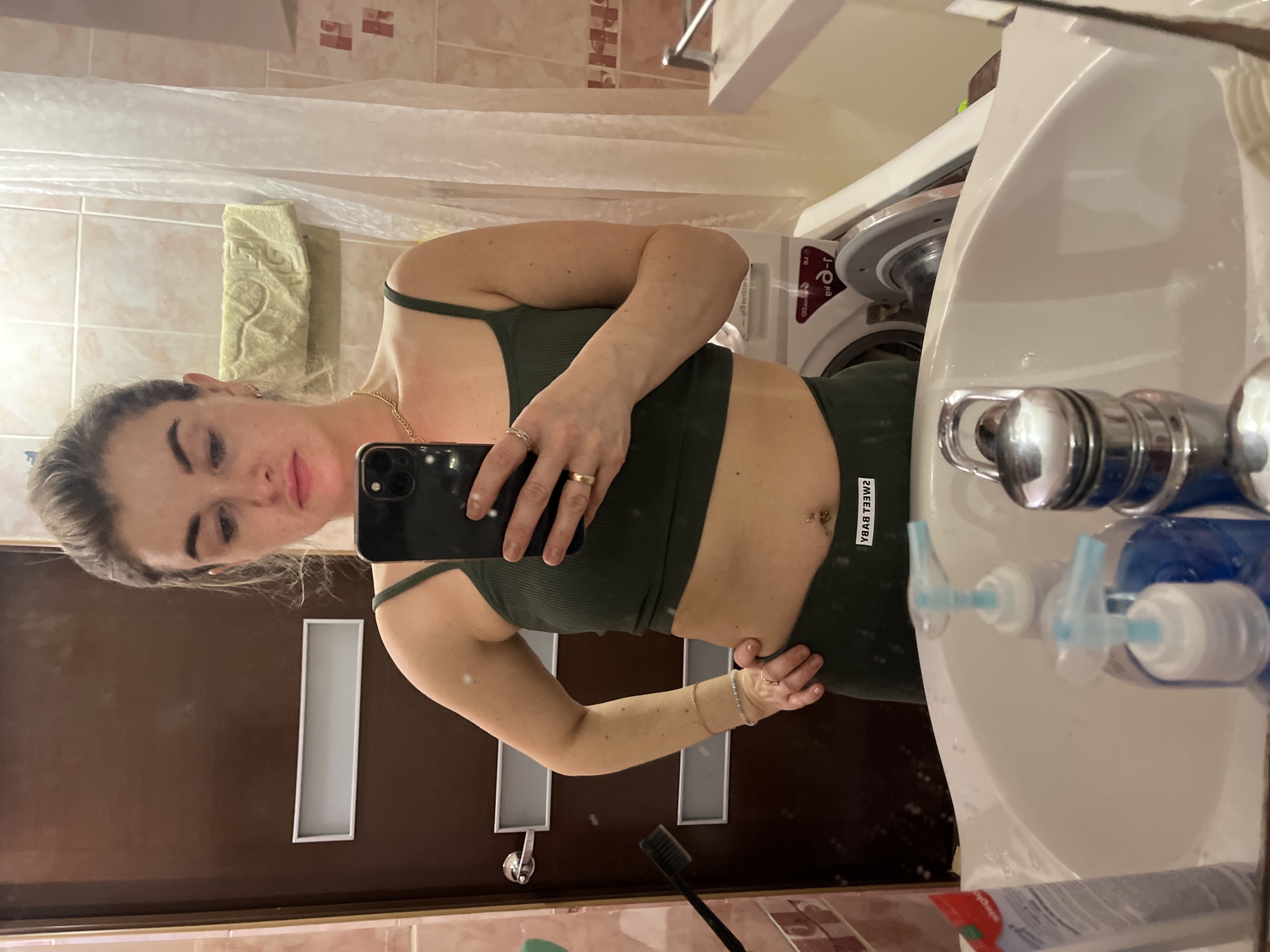 A woman is taking a selfie in a bathroom. She is wearing a green crop top and leggings. The bathroom has a sink, a washing machine, a shower curtain, and a door. A toothbrush, toothpaste, and soap are visible near the sink. The location appears to be a private residence, possibly in an apartment building, but there is no definitive evidence to confirm the specific city of Chaikovsky, Russia.