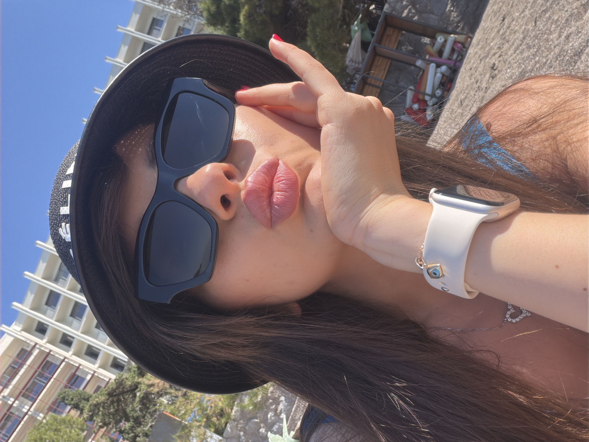 The image is a close-up, diagonally oriented selfie of a person, likely a young adult female, set outdoors in Rafailovići, Montenegro. The subject dominates the frame, wearing a black bucket hat and large, rectangular black sunglasses that cover her eyes. Her long, dark brown hair frames her face and flows down her shoulders.
She is making a playful pose, with her lips puckered as if blowing a kiss. Her left hand is raised to her face, with fingers lightly touching her temple near the sunglasses. Her fingernails are painted bright red. On her left wrist, she wears a white smartwatch with a silver-toned screen, and a small, blue-and-gold charm, possibly an "evil eye," is attached to the watch strap. A delicate silver chain necklace with a small, sparkly heart-shaped pendant is visible around her neck.
The background features a vibrant blue sky and tall, light-colored multi-story buildings with balconies, indicative of an apartment complex or hotel, consistent with a coastal resort area. Some green trees and foliage are also visible. In the upper right, there is a rack holding various colorful cylindrical and rectangular items, which could be beach equipment or other outdoor supplies. The strong, direct sunlight and clear sky suggest a bright, sunny day. The overall impression is one of leisure and vacation. No readable text is clearly visible on any items.