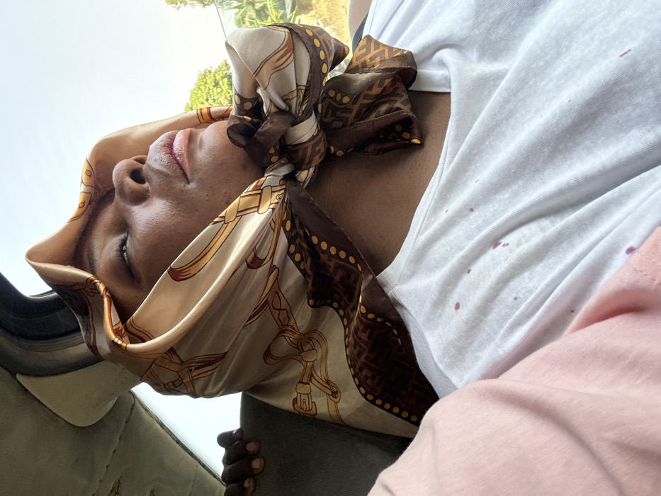 The image depicts an interior scene, likely within a vehicle, during daylight hours, in the city of Mayo, Nigeria. A dark-skinned individual, seen from the mid-chest upward, is the primary subject. Their head is tilted slightly upward, and their eyes are closed or gazing skyward. They wear a light-toned silk headscarf patterned with gold belts and chains, featuring a dark brown border with yellow dots and a repeating 'F' design, tied under the chin. Below the scarf, a white crew-neck t-shirt is visible, marked with multiple scattered reddish-purple stains across the chest area. A pink fabric is partially visible on the immediate left. In the upper left background, the partial hand of another dark-skinned individual is visible. The vehicle's interior headliner, appearing worn, is visible above the hand. Through a window on the right, bright daylight, green foliage, and indistinct background elements are observable.