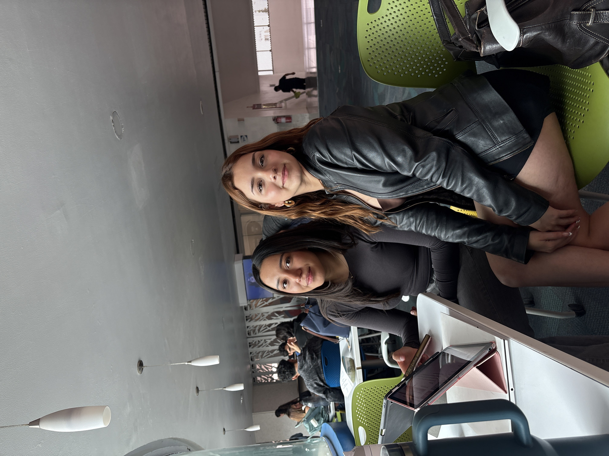 Two young women are seated at a desk with tablets in a public area, likely a library or study space. Additional people are visible in the background seated at desks. The walls are light grey with elongated, pendant light fixtures. Outside the window, a figure is seen walking. The location is in a modern, indoor setting, possibly in Heroica Puebla de Zaragoza, Mexico.
