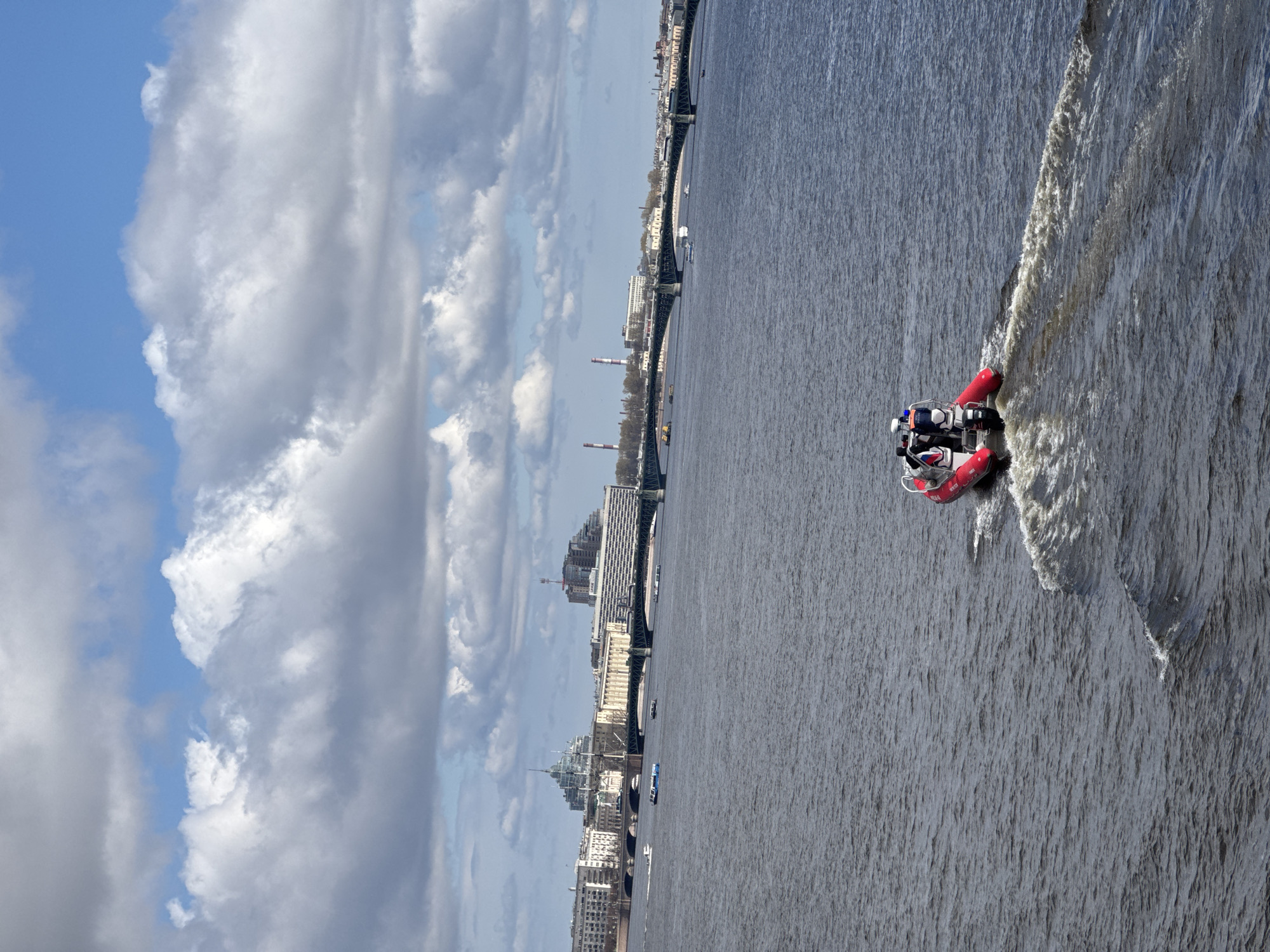 A slightly high-angle shot shows a red inflatable boat traveling on a body of water with the city of Saint Petersburg, Russia in the background, under a cloudy sky.
The inflatable boat is red with white and black details. Two individuals wearing dark clothing and white helmets are in the boat. The boat leaves a foamy wake.
The backdrop reveals the cityscape of St. Petersburg, Russia. Multiple buildings of varied heights and styles can be seen, along with smokestacks and a bridge. The sky is a mix of blue and grey, with mostly large, white cumulus clouds.