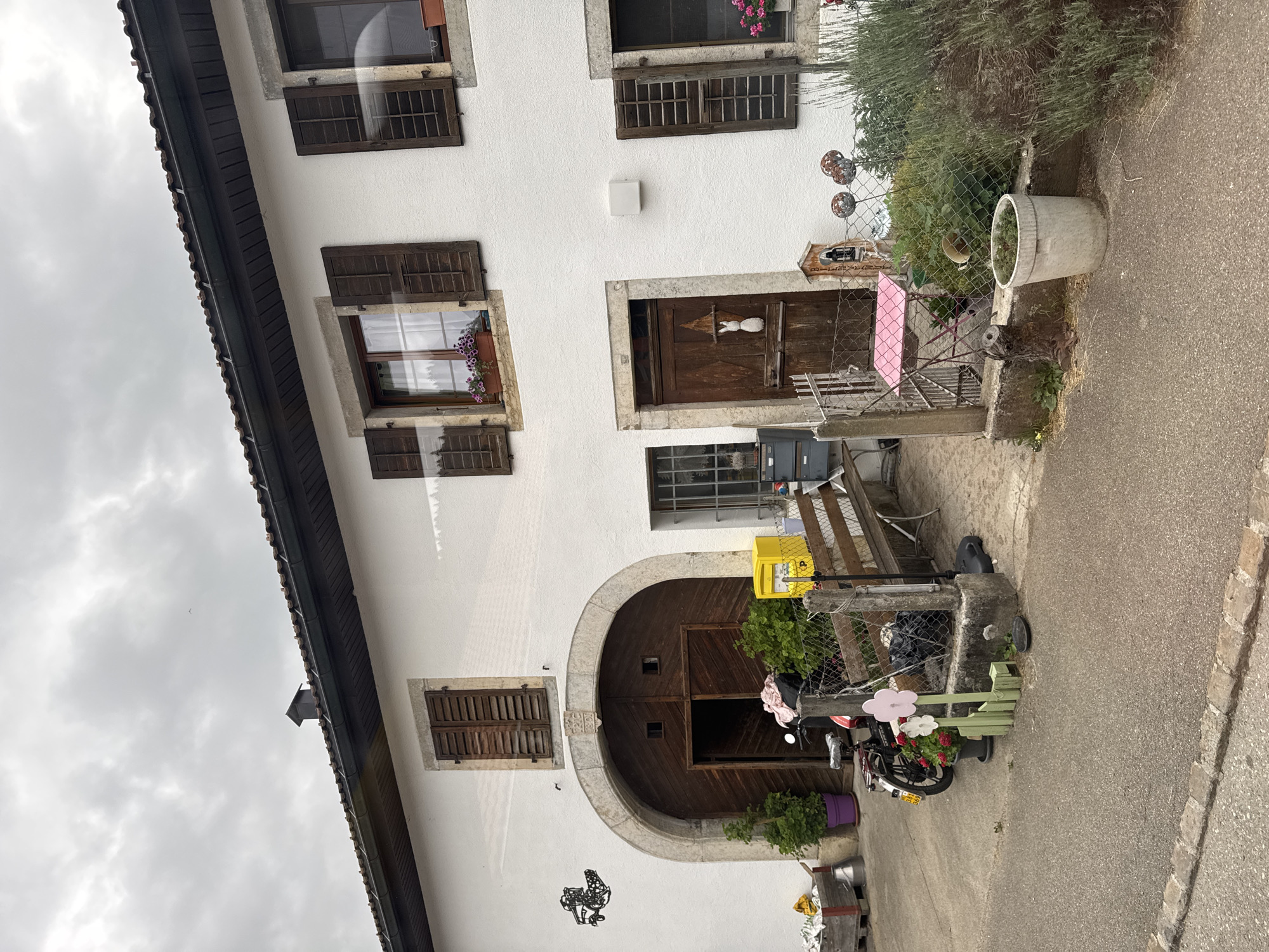 The image shows a building with a white facade and a dark tiled roof. There are several wooden-shuttered windows on the upper floor. A large arched wooden door dominates the left side of the building. To the right of the arched door, there is another wooden door with a white rabbit decoration. A motorcycle is parked in front of the arched door. In front of the building, there is a fence, a wooden bench, and various decorative items including wooden flowers and planters. A yellow mailbox is also visible. The ground is paved with asphalt. The environment appears to be rural, with some vegetation and greenery visible. The image is taken from an eye-level perspective, looking towards the building. The lighting suggests an overcast day.