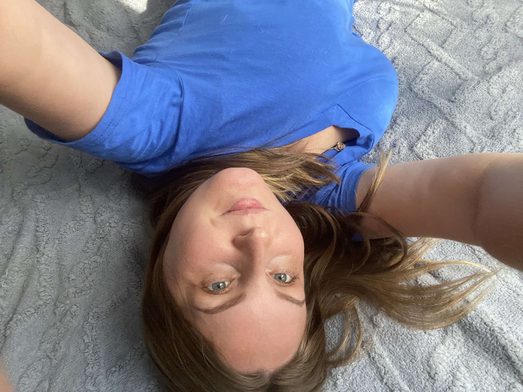 The image shows a young woman with light brown hair and blue eyes lying on a grey, textured blanket. She is wearing a bright blue V-neck t-shirt and a gold necklace with a pendant. Her right arm is extended upwards, and her left arm is bent, with her hand resting on her chest. The lighting suggests it is daytime, with shadows cast on the blanket. The image is taken from a high angle, looking down at the woman. The setting is indoors, with no visible windows or doors. There is no information in the image that can determine the location within the city of Noviy, Russia.