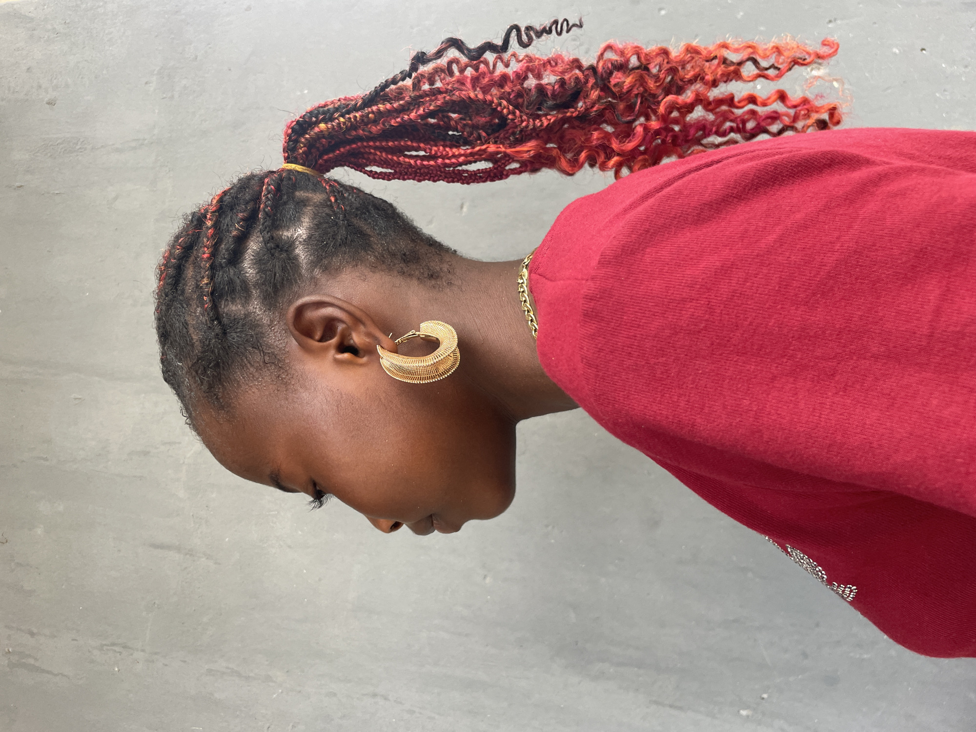 This horizontally oriented image captures a side profile of a person with dark skin, looking downwards. Their hair is styled into a high ponytail, composed of black braids interwoven with distinctive reddish-orange hair extensions. Some parts of the extensions are curly, adding texture to the hairstyle, which is secured with a yellow hair tie.
The visible ear is adorned with a prominent, intricate golden hoop earring, featuring a layered, ridged design. A delicate gold chain is visible around the person's neck. They are wearing a plain red long-sleeved garment, which appears to be made of a soft fabric like cotton. Towards the bottom right of the red garment, there are small, shiny silver embellishments, possibly beads or glitter.
The background consists of a plain, light gray, textured wall or surface, providing a neutral backdrop that highlights the subject. The lighting is bright and even, suggesting it is daytime. The person is in a static pose, with no specific activity taking place. No text is visible within the image. The context provided indicates this image was taken in Badagry, Nigeria.