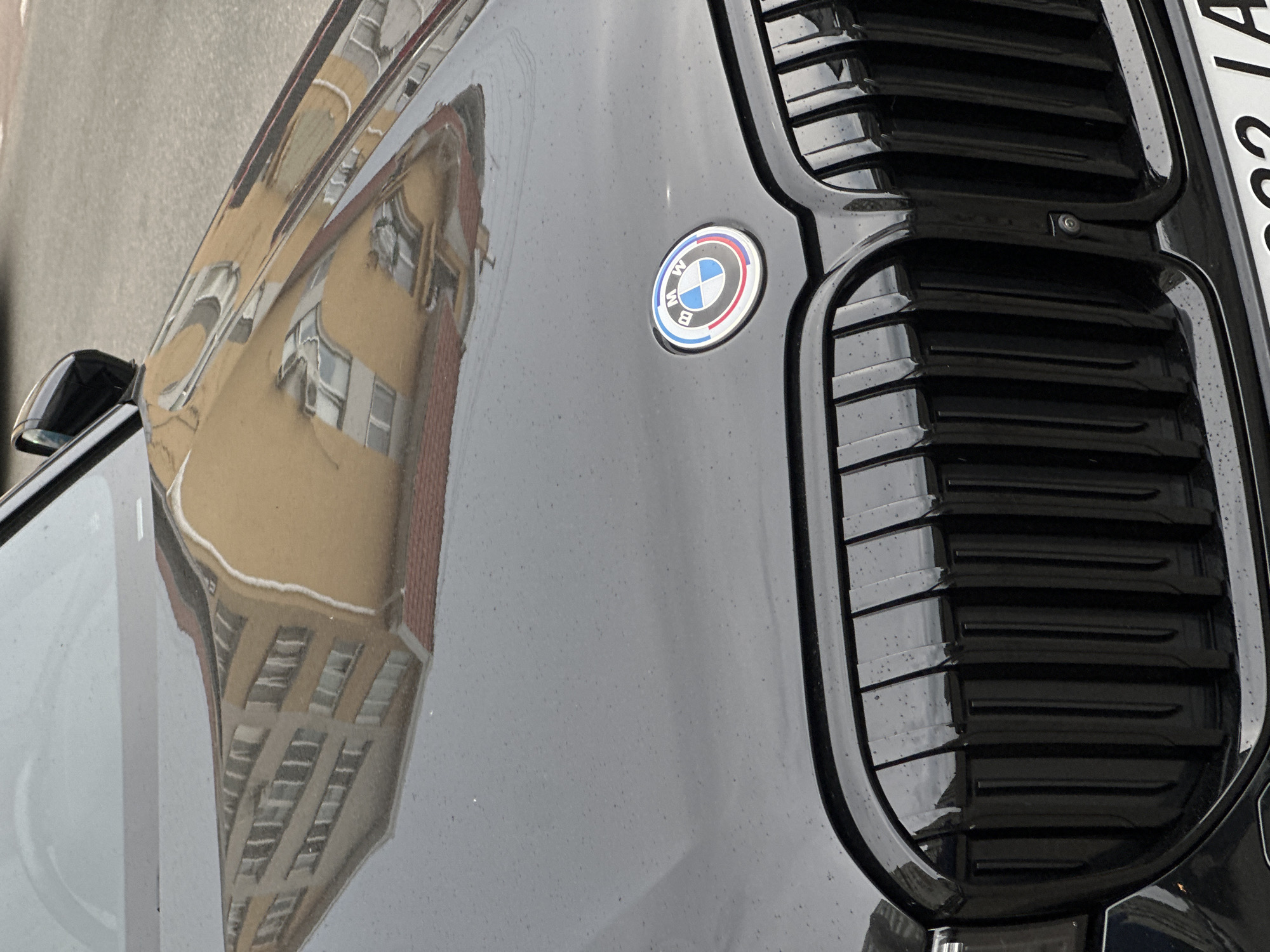 The image shows the front grille and hood of a black BMW. The BMW logo is visible on the hood, centrally located above the grille. The grille itself is black and has a vertical slat design. The hood is also black and shows reflections of a multi-story building with many windows and a reddish-brown roof, as well as a side-view mirror and part of a street. There are small water droplets on the car's surface, suggesting recent rain or dew. The lower part of the image shows a portion of a license plate with white text on a black background. The environment appears to be an urban setting, as indicated by the building's reflection. The location is specified as Kriukivshchyna, Ukraine. There are no people or animals visible in the image.