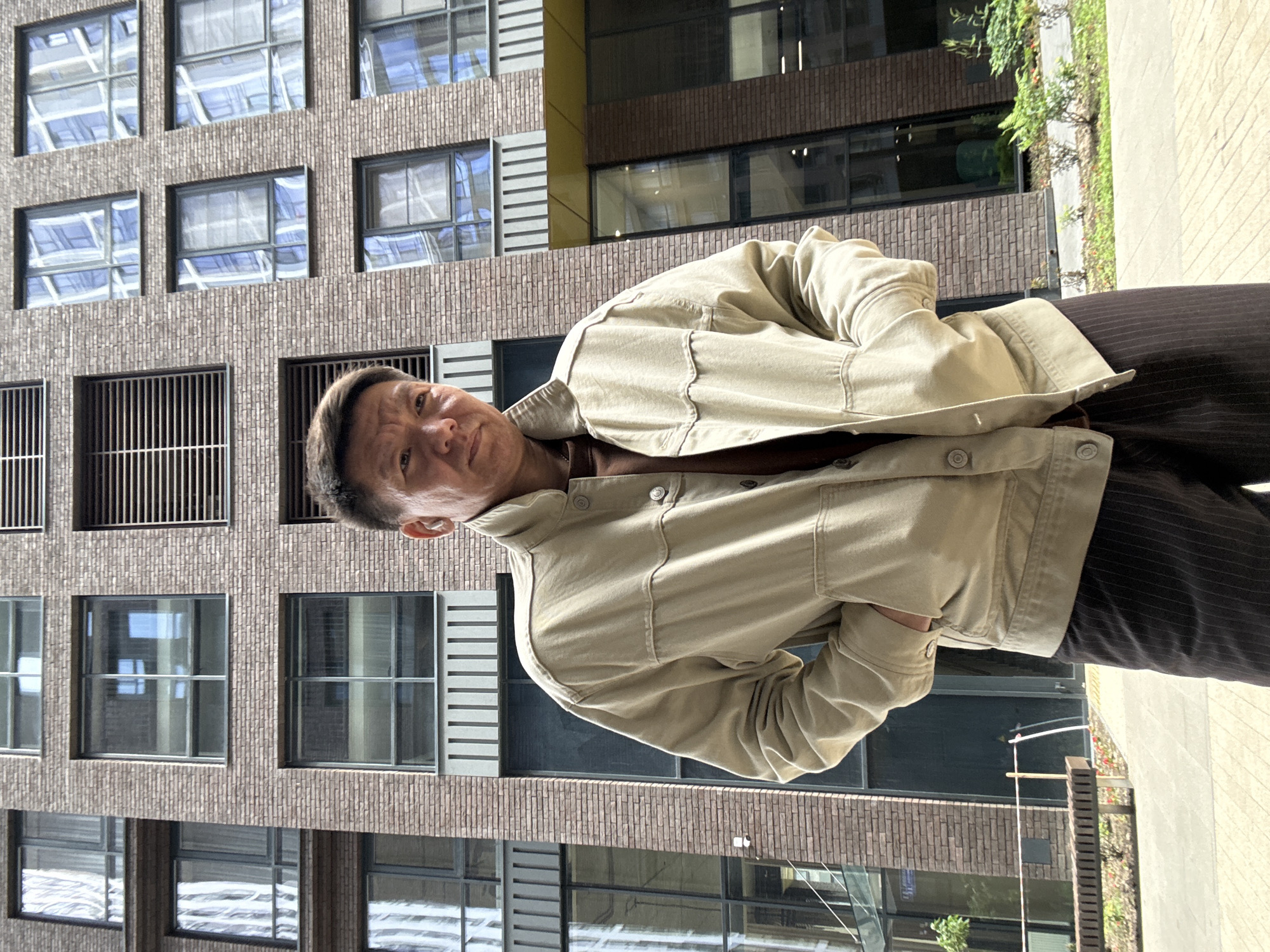 The media file is a portrait of a man with short dark hair, wearing a light tan jacket over a brown shirt and dark pinstripe pants. He is leaning to his right, with his head tilted slightly upwards and his gaze directed towards the camera. One hand is placed in the pocket of his jacket.
The man is positioned in front of a modern brick building with numerous windows. The windows reflect the sky and the surrounding environment, suggesting it is daytime. The building's architecture features a textured brick facade and horizontal window frames. Greenery, possibly a vertical garden or trees, is visible on the right side of the frame, adding a natural element to the urban setting.
The scene appears to be outdoors on a sunny day, indicated by the bright lighting and shadows. The man's expression is neutral, and the overall mood is casual and relaxed. The location is implied to be Moscow, Russia, based on the provided context. There is no visible text in the image.