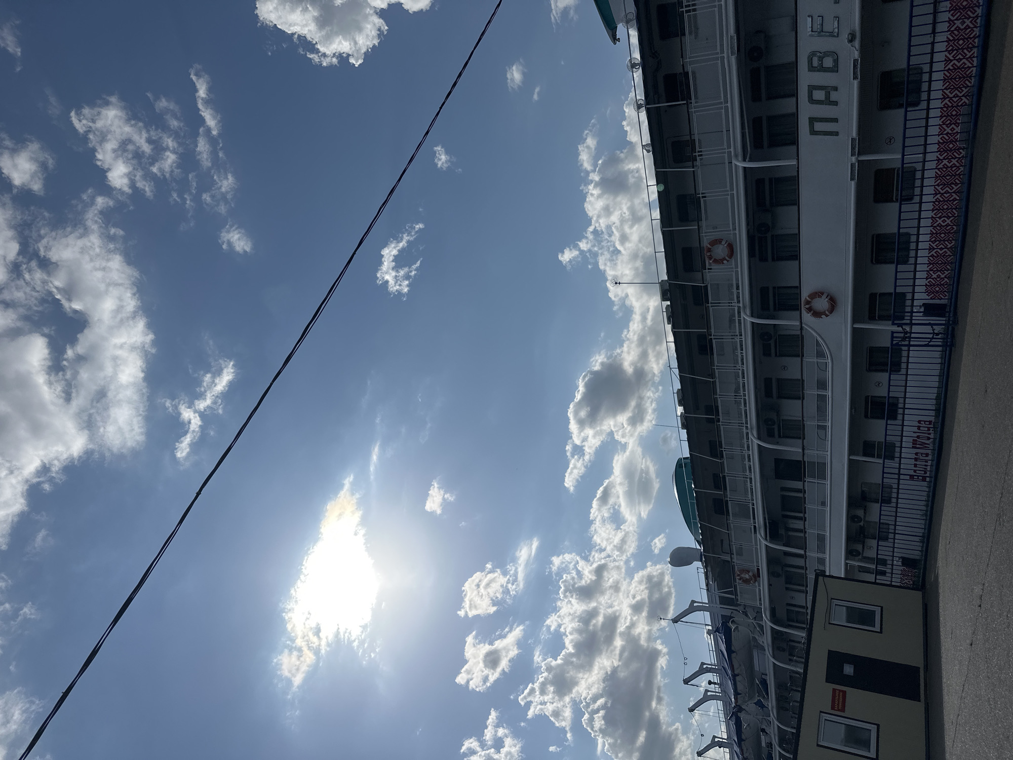 The image depicts a partially visible cruise ship docked at a pier, against a backdrop of a blue sky with scattered cumulus clouds. A power line stretches diagonally across the upper left portion of the frame, intersecting the sky. The sun is partially obscured by clouds, creating a bright, diffused light. The side of the ship, displaying the name "ПАВЕЛ" in Cyrillic script, features multiple decks with windows and life preservers. A small, unidentifiable building adjoins the ship. Based on the writing visible on the boat "Yedkina Volga", the location is determined to be the city of Chaikovsky, Russia, as a boat cruise company with that name operates there.