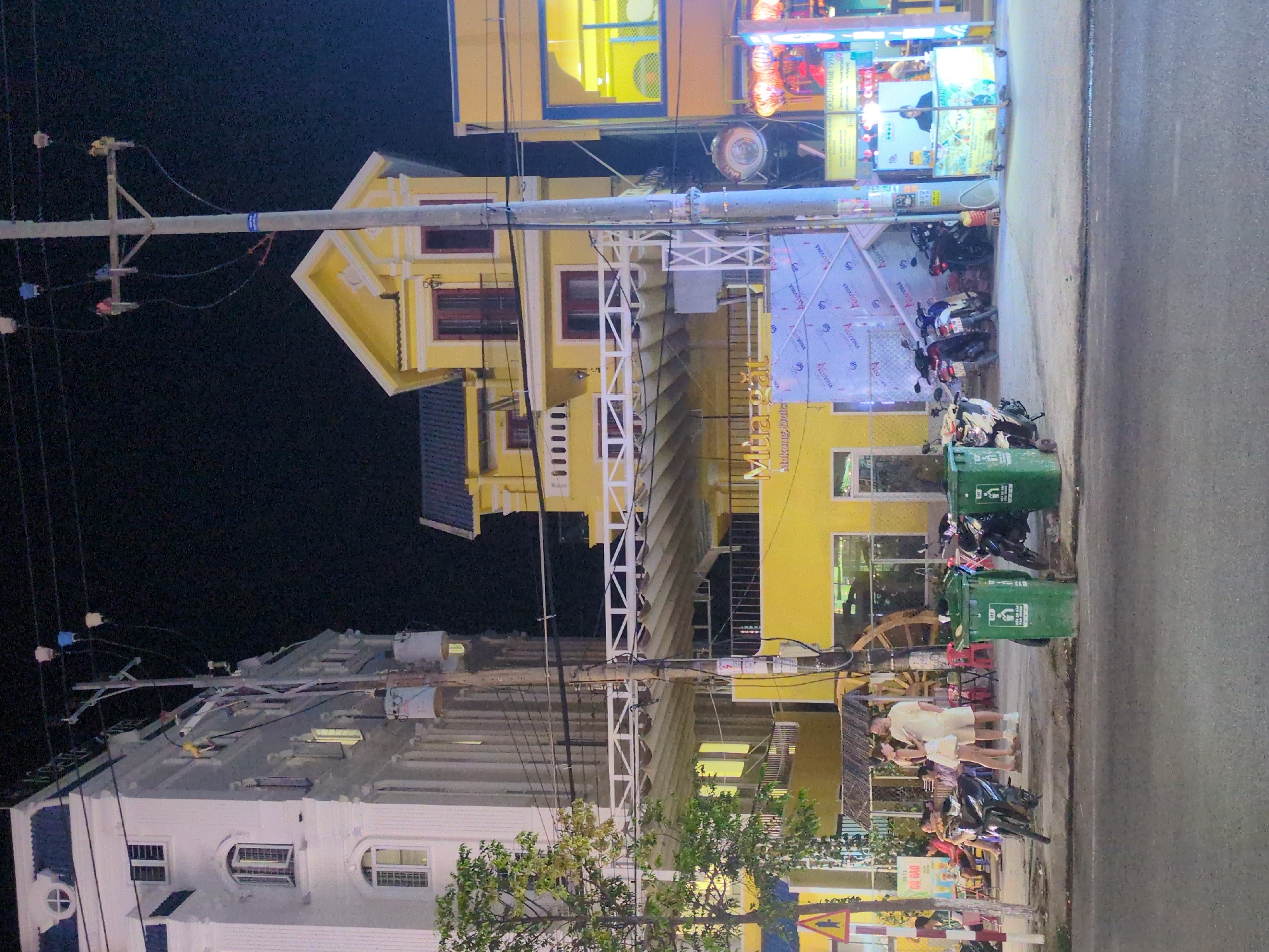 Here's a detailed description of the image:
**General Scene:** The image captures a street scene at night in Phú Quốc, Vietnam. The street is lined with buildings and lit by streetlights and neon signs.
**Buildings:** There are two prominent buildings: a white, two-story structure on the left and a taller, yellow building on the right. The white building has a classical architectural style, with arched windows. The yellow building has a more modern design.
**Objects:** Utility poles with wires run along the street. Several parked motorbikes are visible. Green trash cans are located on the sidewalk. Various signs and advertisements are displayed along the buildings.
**People:** A small group of people are standing on the sidewalk, engaged in conversation.
**Action:** People appear to be standing and interacting in front of a shop or restaurant. Motorbikes are parked, suggesting people may be visiting the establishments.
**Environment:** The scene suggests a commercial or residential area with a mix of older and newer buildings. The presence of motorbikes and pedestrians indicates a busy urban environment. The time of day is night.