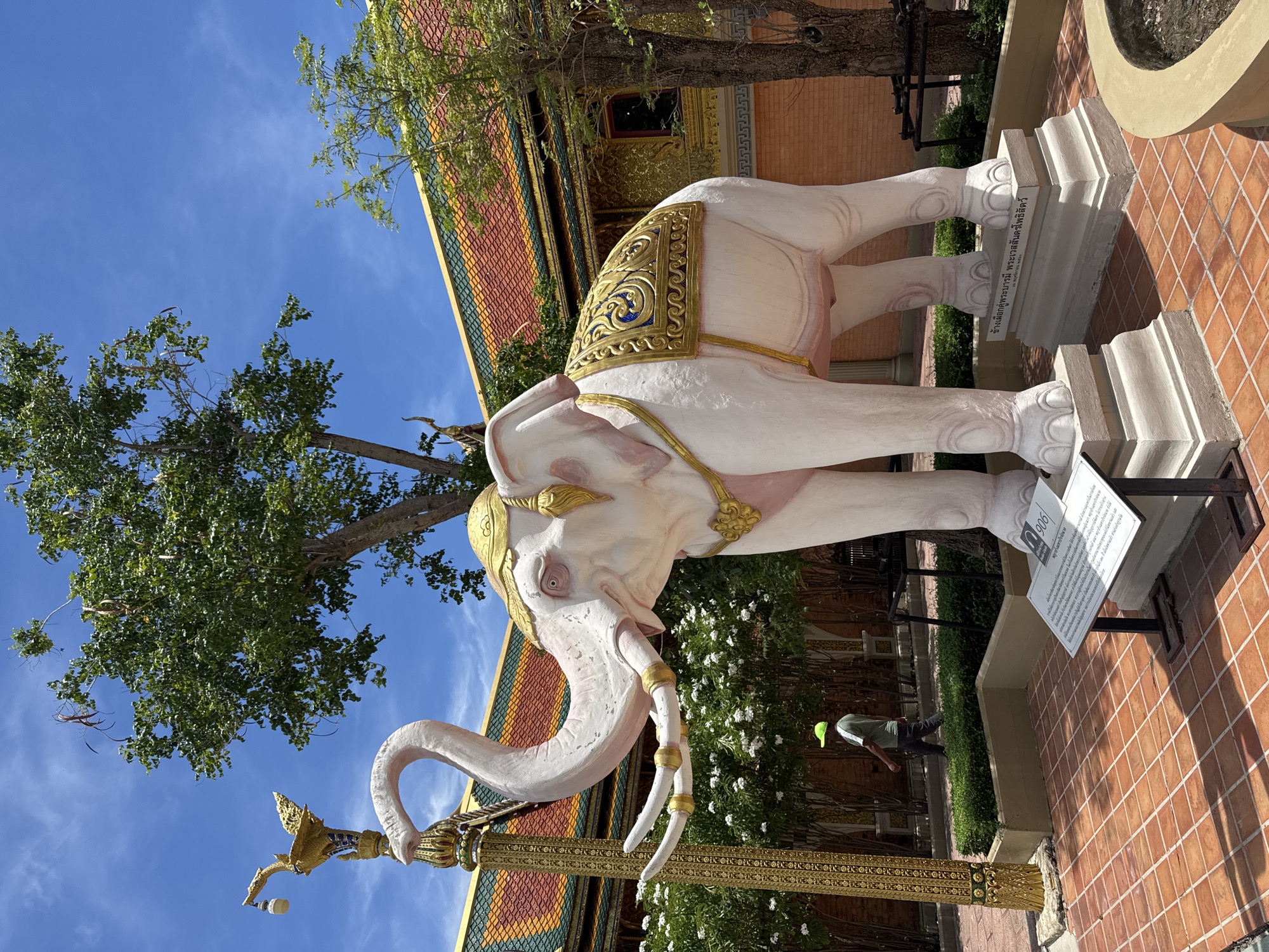 A low-angle shot depicts a white elephant statue with golden embellishments standing outside a building with a multi-colored roof in Bang Pu Subdistrict Municipality, Thailand. The elephant stands on a square stone base with signage and a smaller white sign in front. A golden pole with a bird-like sculpture is to the left of the elephant. A tree with green leaves is situated behind the elephant, set against a blue sky with thin clouds. In the background, behind the elephant, there is a view of landscaping with white flowers, along with a person wearing a green cap and a dark shirt and pants. The ground around the statue is covered in red tiles.