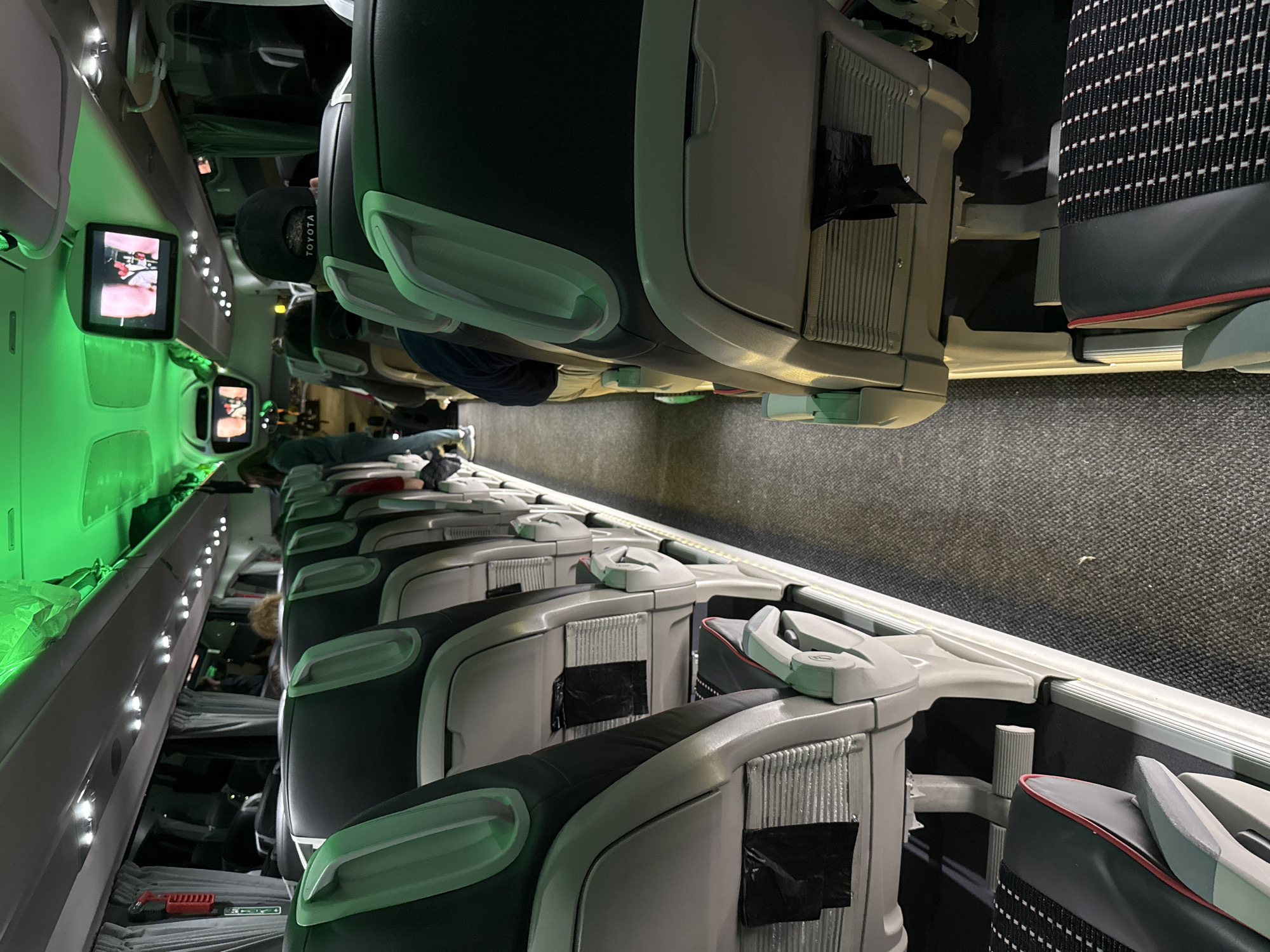 The image shows the interior of a commercial airplane cabin. Rows of seats with headrests are visible on both sides of the aisle. The seats are dark gray with light gray accents. Some seats have black coverings over the in-flight entertainment screens. Passengers are seated in some of the rows. Overhead compartments line the ceiling, with a green-lit panel running along the side of the plane. The lighting in the cabin is dim. The floor is carpeted. A passenger is wearing a hat with the word "TOYOTA" on it. There is no indication that the image is related to the city of Veliko Tarnovo, Bulgaria.