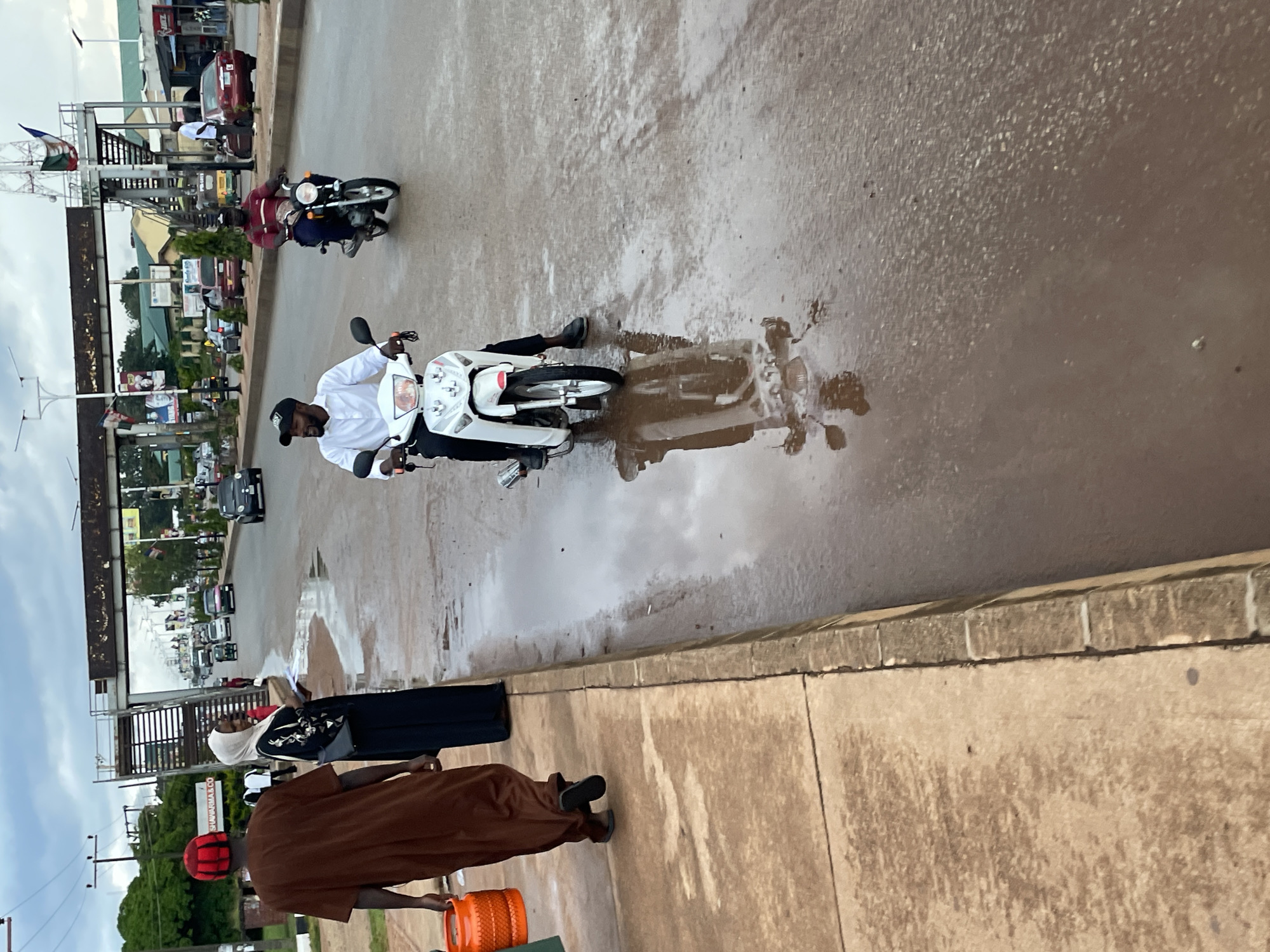 The image depicts a street scene in Jos, Nigeria, likely after a rainfall, as evidenced by the standing water and reflections on the asphalt. In the foreground, a person wearing a brown traditional outfit and a red kufi is walking on the sidewalk, carrying an orange plastic container. Beside them, a woman in a black hijab and abaya stands with a piece of paper. A busy street is to their right, with several vehicles including cars, a tricycle, and motorcycles. One motorcycle rider, dressed in a white shirt and black pants, is prominently featured in the middle of the road, his reflection visible in the wet surface. Another motorcycle rider in a red shirt is further down the road. Overhead, a pedestrian bridge spans the street. The background shows commercial buildings, trees, and utility poles, contributing to the urban setting.