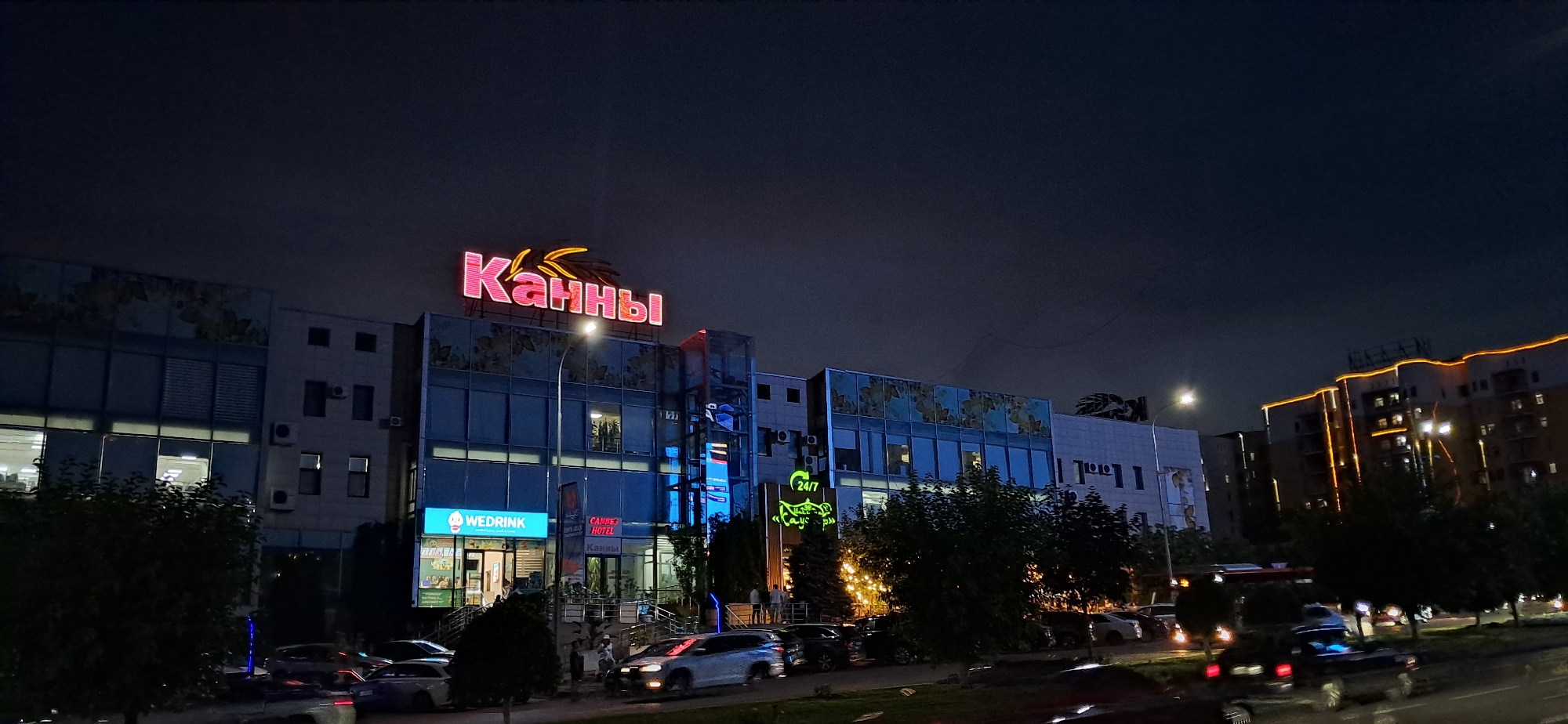 This image shows a nighttime street view of a multi-story building complex. A large, neon pink sign that reads "КАННЫ" is prominently displayed on the roof of the central building, with a stylized image of wheat stalks above the lettering. Below this sign, the building features large glass windows and a glass-enclosed elevator shaft illuminated with blue lights. Several signs are visible on the lower floors, including "WEDRINK", "CANNES HOTEL", and "24/7". The building is illuminated by streetlights and interior lights, and the facade is decorated with blue accents and what appear to be floral patterns.
To the right of the central building, another building is visible, also illuminated by external lights along its edges and windows. Trees and bushes are present along the sidewalk and in front of the buildings. Several cars are parked along the street and driving on the road in the foreground. The sky is dark and appears overcast.