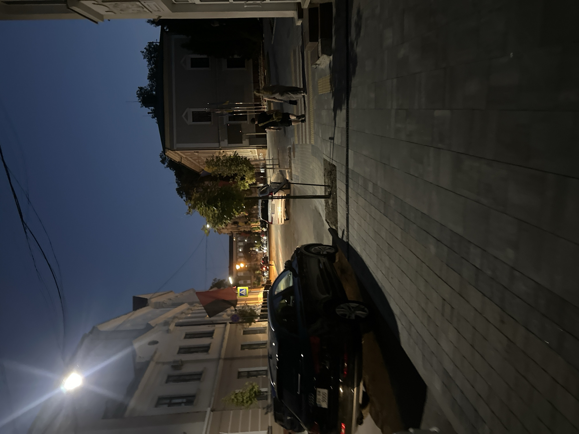 An evening scene on a city street in Ternopil, Ukraine, shows a paved sidewalk and street illuminated by streetlights. On the sidewalk, two individuals are walking away from the camera, a man with a bag slung over his shoulder and a woman. In the street, a black Audi SUV is parked, and further down, another car is visible. Buildings with windows line the street on the left and right. A flag, red and black, hangs from a pole on a building to the left. The sky is a deep blue, indicating dusk or early night.