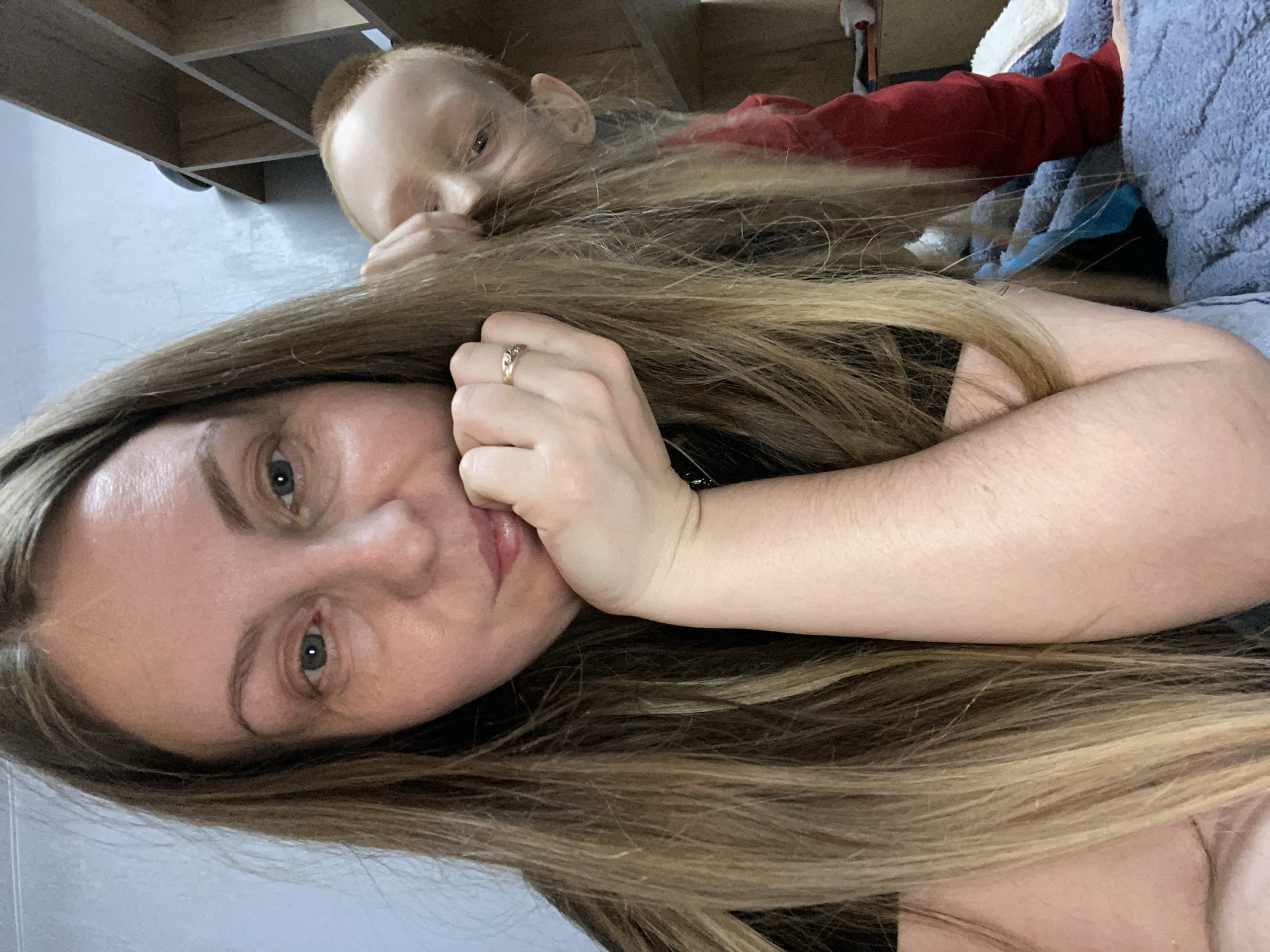 A woman with long blonde hair, a gold ring on her ring finger, and blue eyes is lying down. A child with fair skin and red hair is lying behind her, partially obscured by her hair. A shelf and a red shirt are visible in the background. A gray blanket or towel is also present in the background. The location is not discernible from the image.