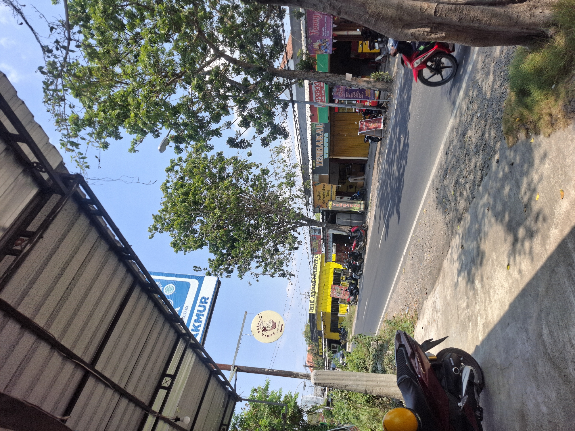 This image depicts a street scene in Banjarmadu, Indonesia, under a clear blue sky. The foreground shows a concrete sidewalk with dappled sunlight and shadow. A parked maroon scooter with a yellow helmet is visible on the left. The street itself is paved and appears to be a two-lane road with some wear and tear. Several motorcycles are parked along the side of the road in front of businesses.

On the right side of the street, a row of shops with awnings lines the road. One shop has a yellow facade with "MIE AYAM MARNYU" written on it. Another shop displays a sign for "TEMBAKAU WILOWO". Further down, a shop with a purple and pink banner reads "Funny". Trees with green foliage are scattered throughout the scene, with one large tree trunk prominently featured on the far right. A street lamp is visible in the distance, mounted on a pole. The overall impression is of a commercial area in a small Indonesian town.