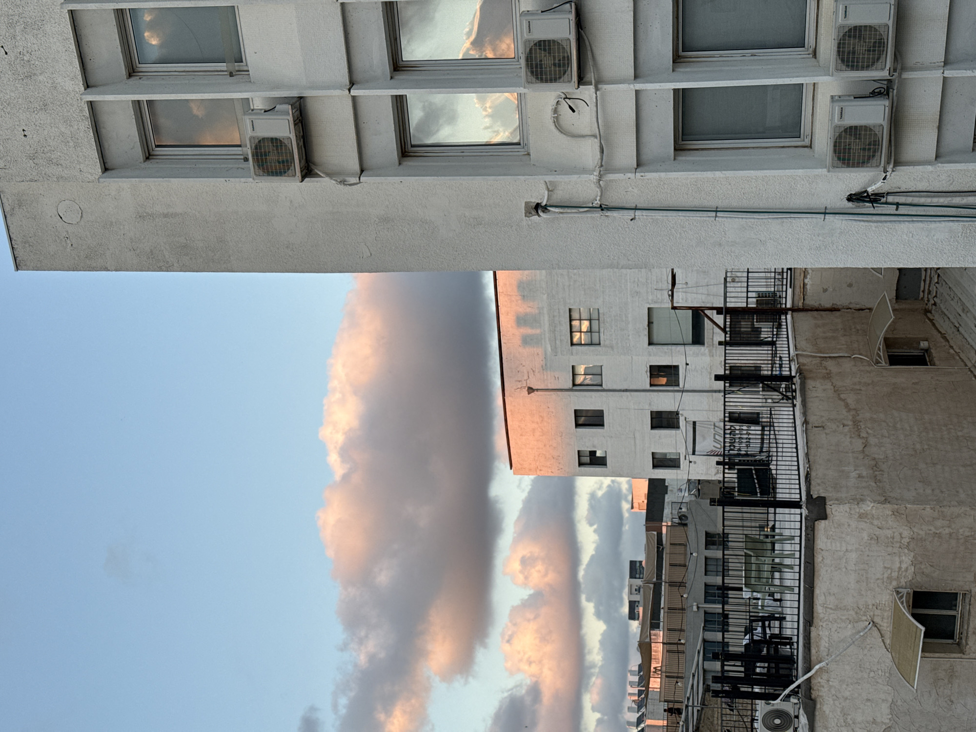 The image shows an exterior view of buildings in Tel Aviv, Israel, with a sky containing clouds reflecting sunset colors. The foreground building is white with multiple windows, some reflecting the sky, and air conditioning units attached. A second building, also white, is visible behind it, also with windows and balconies with black railings. The buildings are of a typical architectural style for Tel Aviv. The sky is blue with large, pink-tinged clouds. Utility lines and other building infrastructure are visible along the walls.