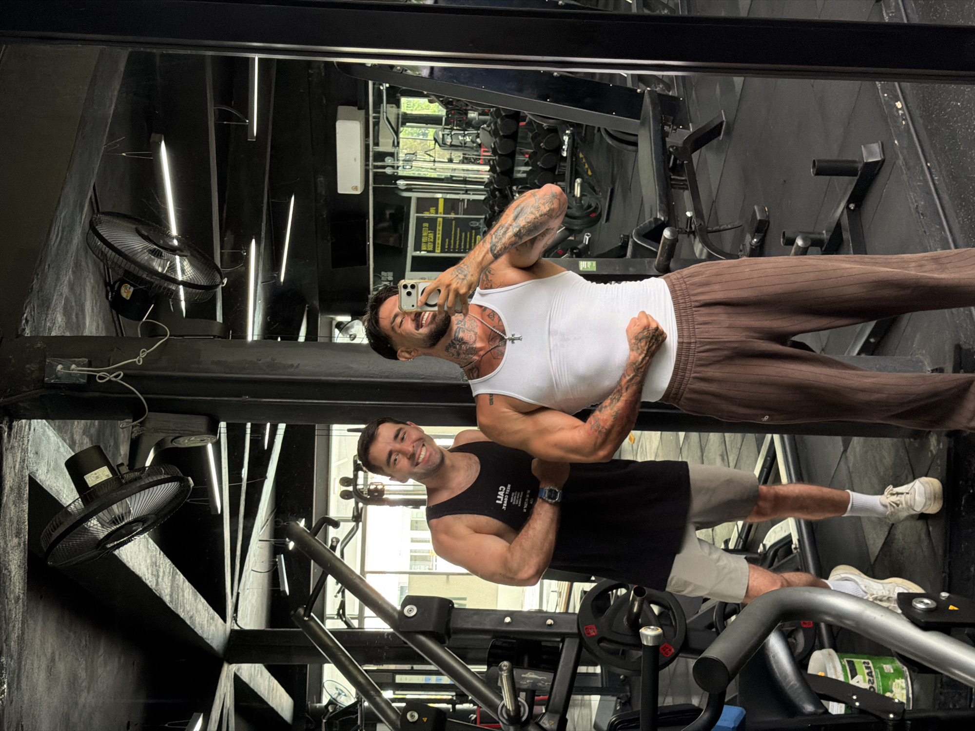 Two men are posing in a gym, likely taking a selfie in a mirror. The man in the foreground is wearing a black tank top and light-colored shorts with white socks and sneakers. The man reflected in the mirror is wearing a white tank top and brown pants. Gym equipment, including weight machines and dumbbells, can be seen in the background. Ceiling fans are visible, and the overall lighting is bright. The location is Kathu, Thailand, as indicated by visual cues such as the gym's interior design.