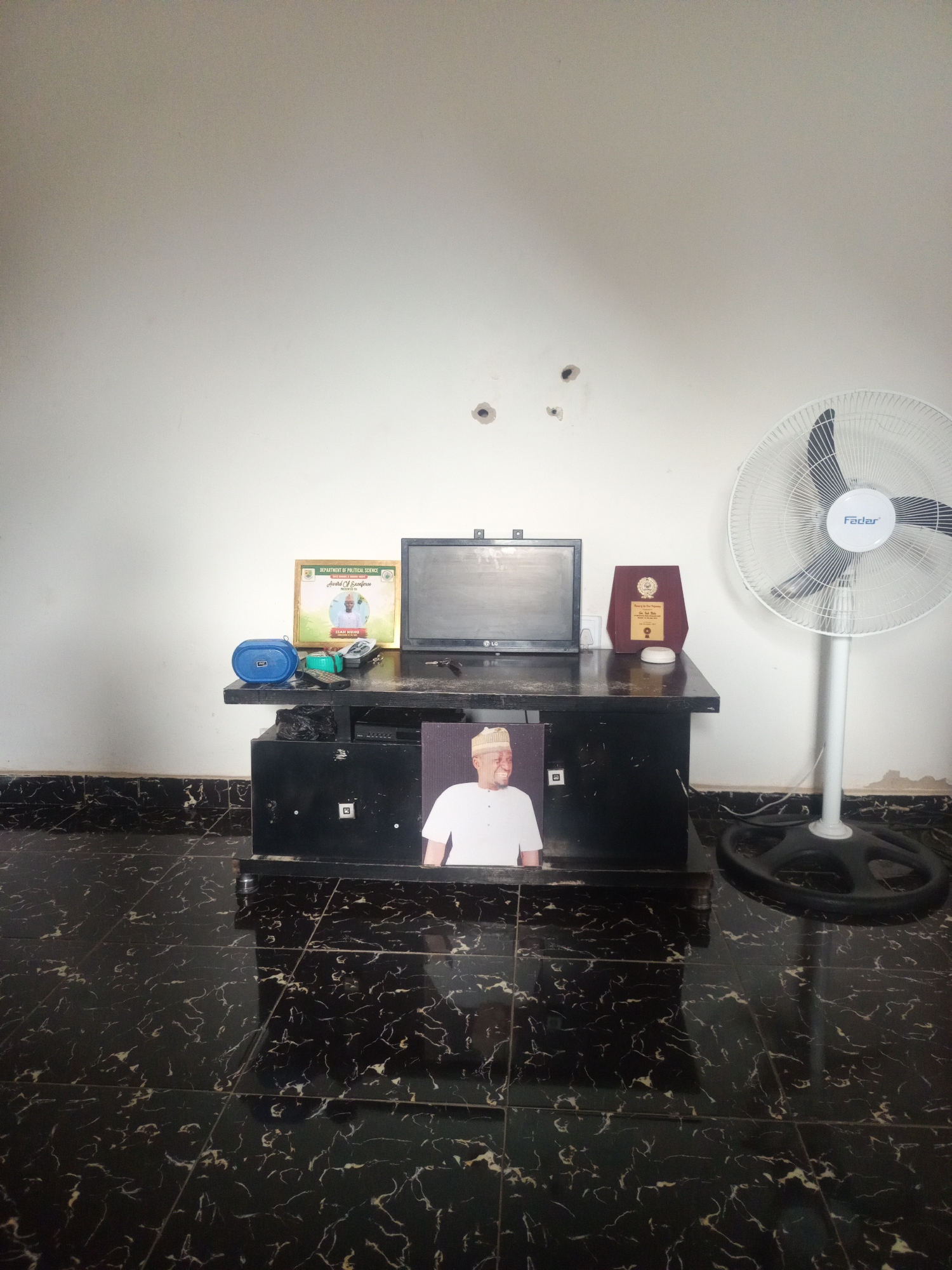 The image displays an indoor scene within a room in Anguwar Jaba, Nigeria. The room features white painted walls, exhibiting some minor scuffs and three distinct circular holes in the upper-middle section, suggesting previous wall hangings. The floor is composed of dark, possibly black, square tiles with prominent white and light gold marbling patterns.
A dark-colored, rectangular media console or television stand is centrally positioned. On its top surface, from left to right, are: a blue portable speaker with "JBL" visible on its front grille, several remote controls, a small black LG-branded monitor, and two awards. The award on the left is a framed certificate with green and gold accents, displaying text "DEPARTMENT OF POLITICAL SCIENCE" and "Award Of Recognition," featuring a photograph of a bearded man in a cap, identified by the partial name "ISAH NURU." To the right of the monitor is a dark wooden plaque with a metallic plate. A small white cylindrical object rests in front of the wooden plaque.
Propped against the front base of the media console is a large framed photograph of a smiling man in a white shirt and a light-colored cap, facing slightly to the viewer's left.
To the right of the media console, a white "Fadar" brand pedestal fan stands on the floor, featuring black blades behind a white protective grille. Electrical cables are visible on the floor behind the fan and the media console. The general environment appears lived-in but tidy.