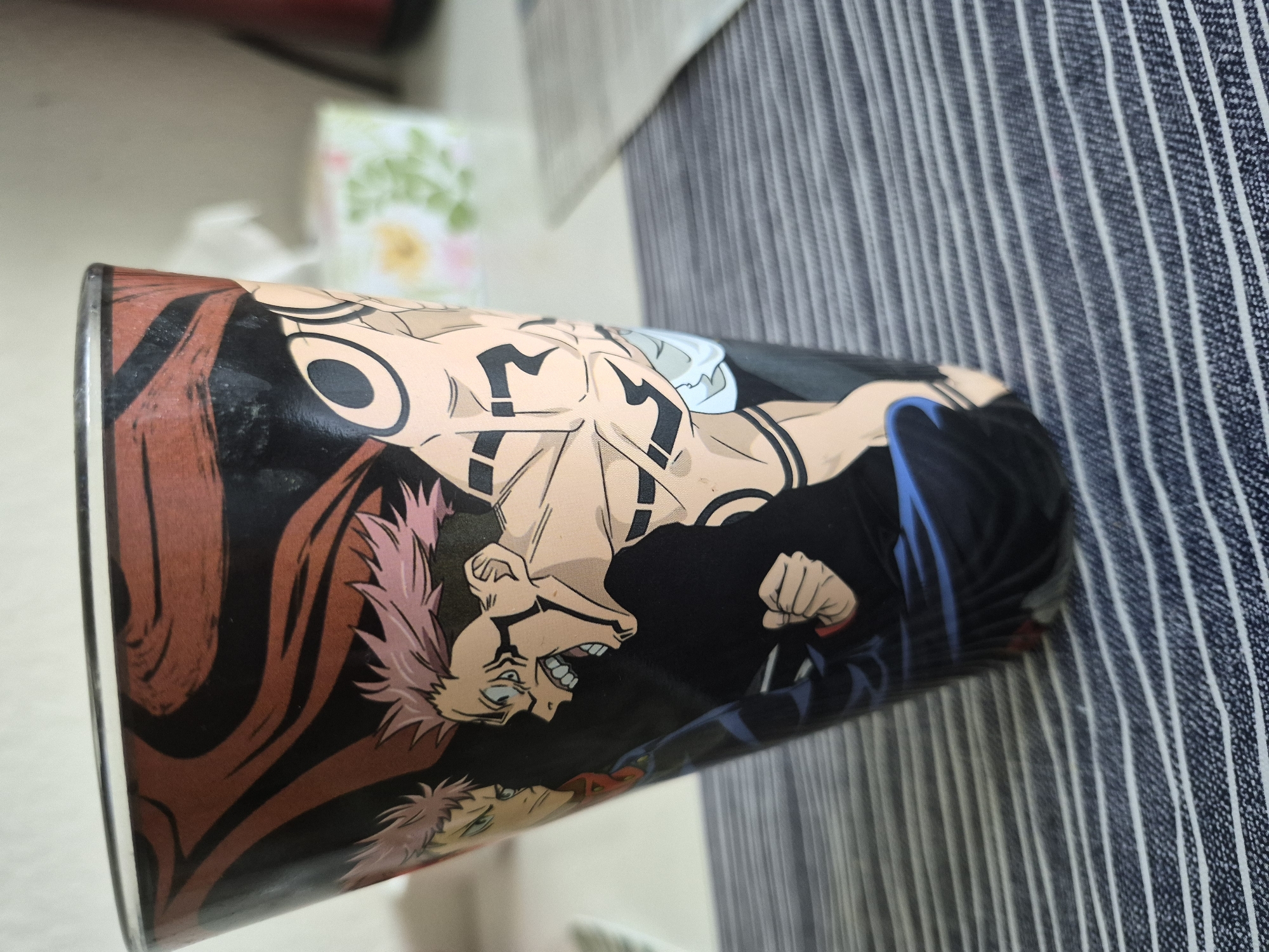 The image shows a close-up, side-view of a tall, conical glass with a colorful anime design. The design features two characters with spiky pink hair, one facing left with a stoic expression and the other facing right with an open-mouthed, aggressive expression. Both characters have distinctive black markings on their bodies, with the character on the right displaying extensive tribal-like tattoos on their torso and arms. The background of the design is black with flowing red and brown brushstrokes at the top. The glass rests on a surface with a striped pattern, appearing to be a placemat or fabric. In the blurred background, out of focus, are some white tissues and a small decorative box with floral patterns. The lighting suggests an indoor setting, possibly a table. The scene does not provide any information about a specific city or location.